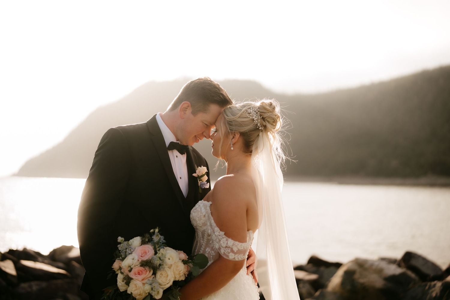 Bride and groom share a romantic moment by rocky shoreline with mountains and golden sunset light in background.