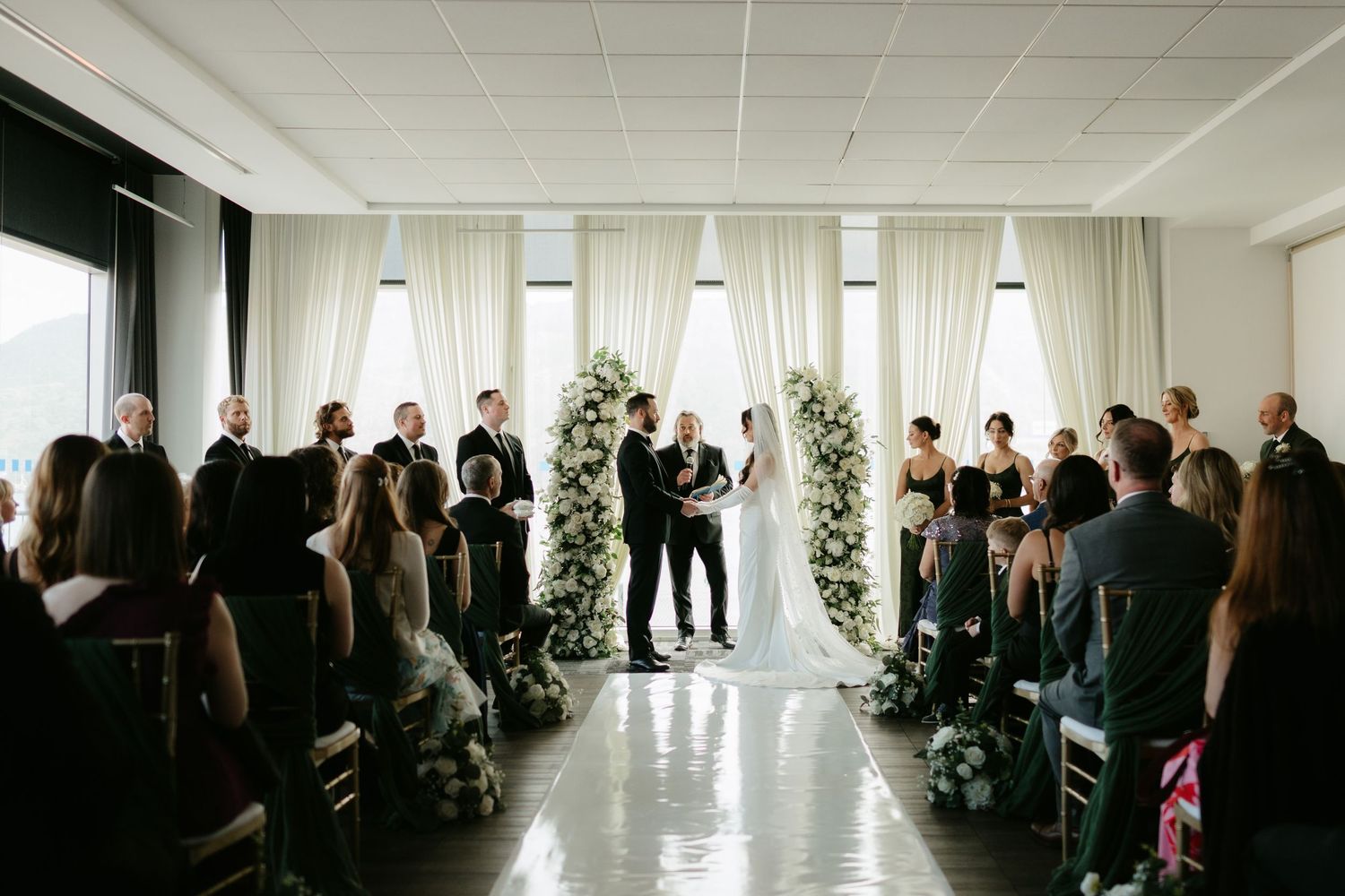 Elegant indoor wedding ceremony with white floral arch, draped curtains, and guests seated in modern venue.