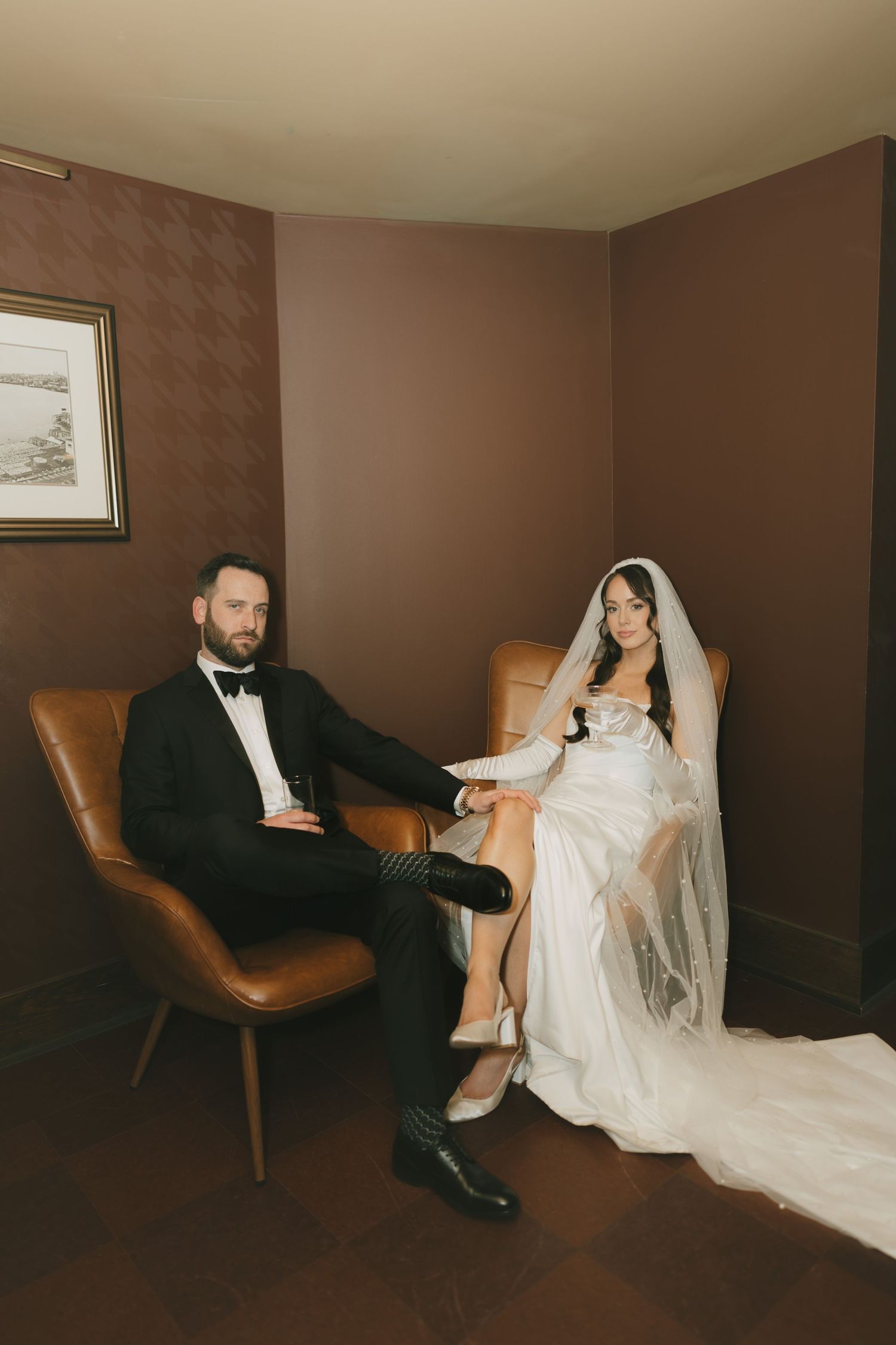 A bride in a white gown and veil sits beside a groom in a black tuxedo on leather chairs in a moody brown room.