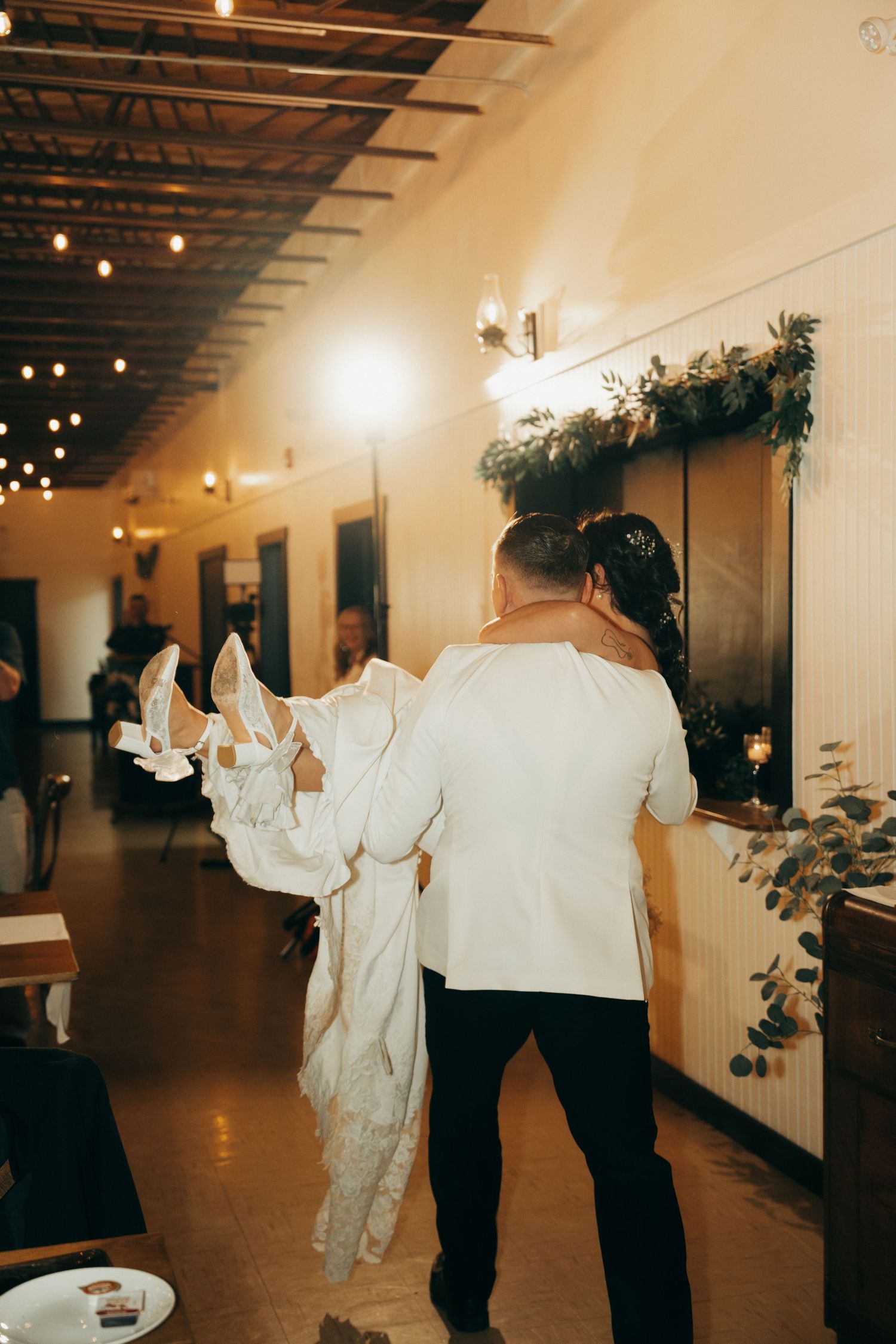 Wedding guests dance together in an indoor venue with warm lighting and greenery decorations on the walls.