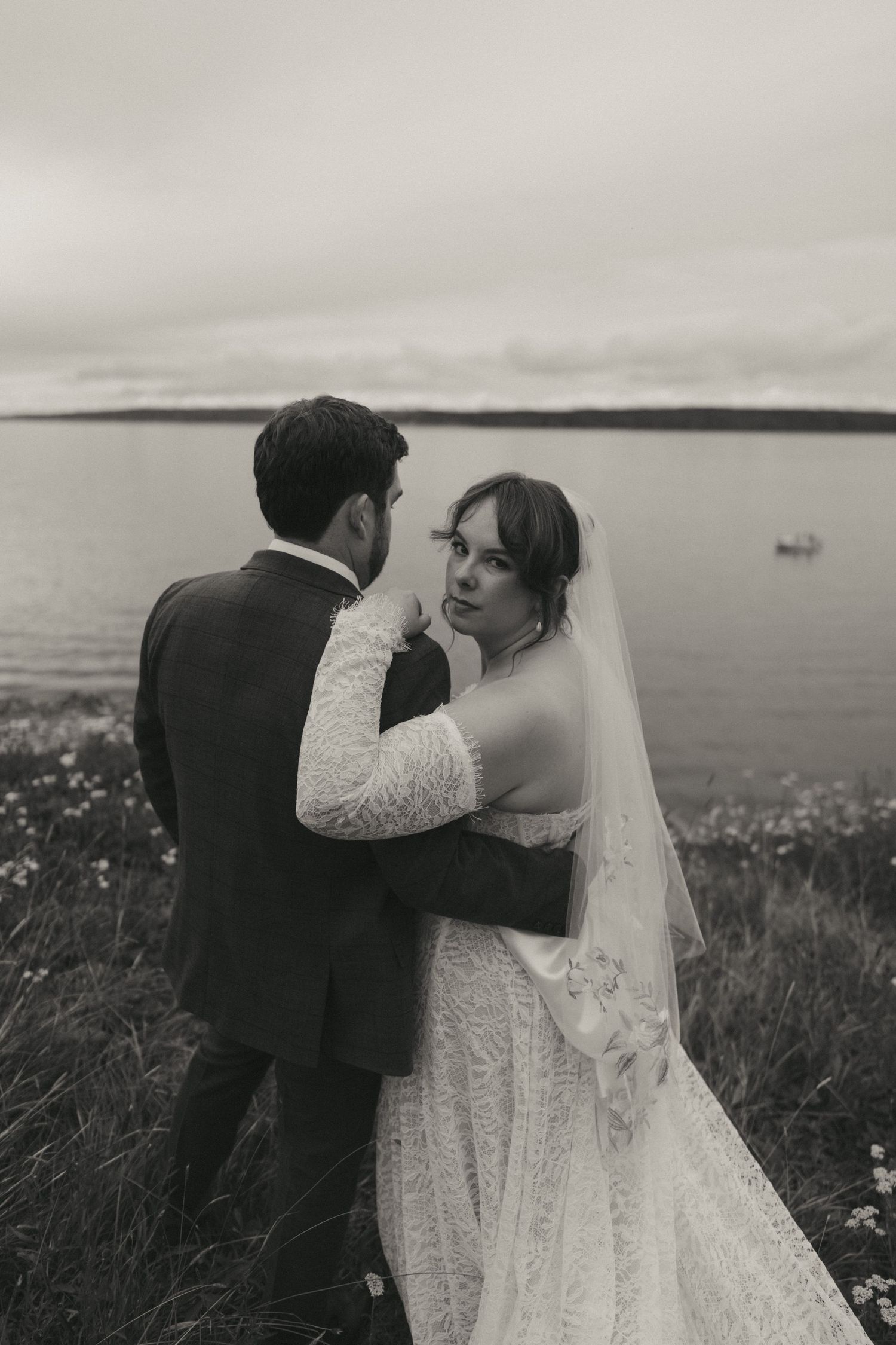 Black and white photo of a bride and groom embracing near a coastal landscape on their wedding day.