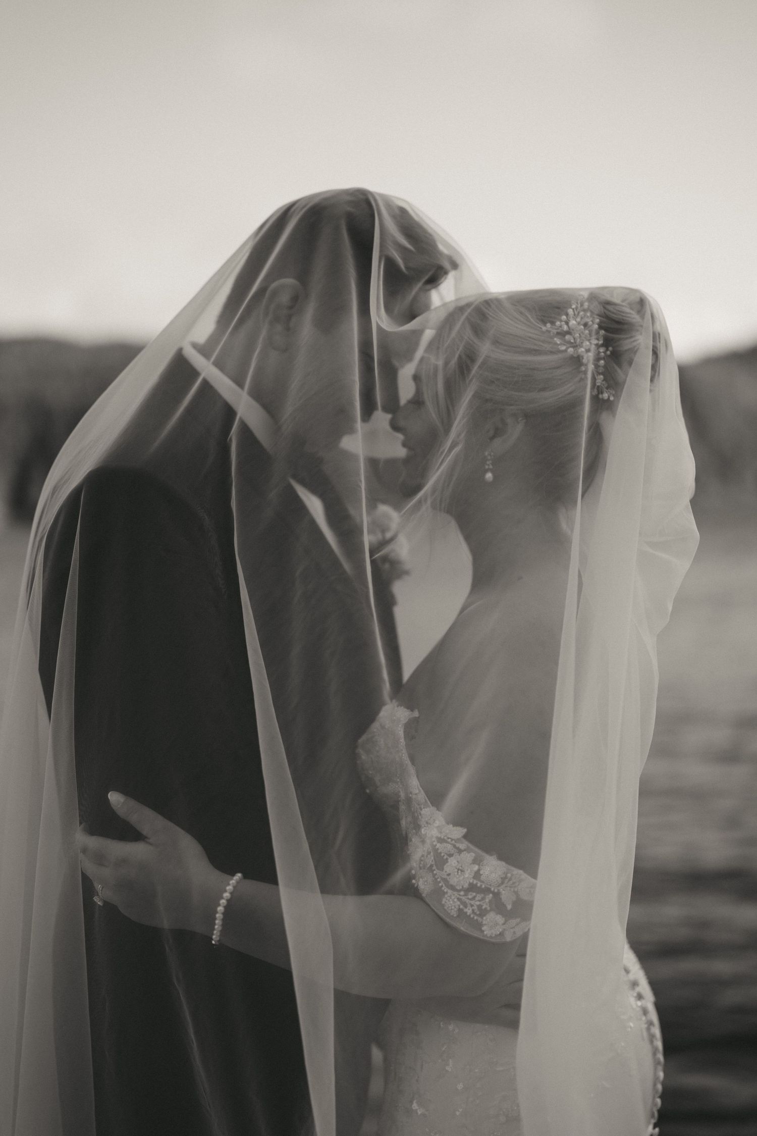 Black and white photo of bride and groom sharing a romantic moment under a flowing wedding veil.