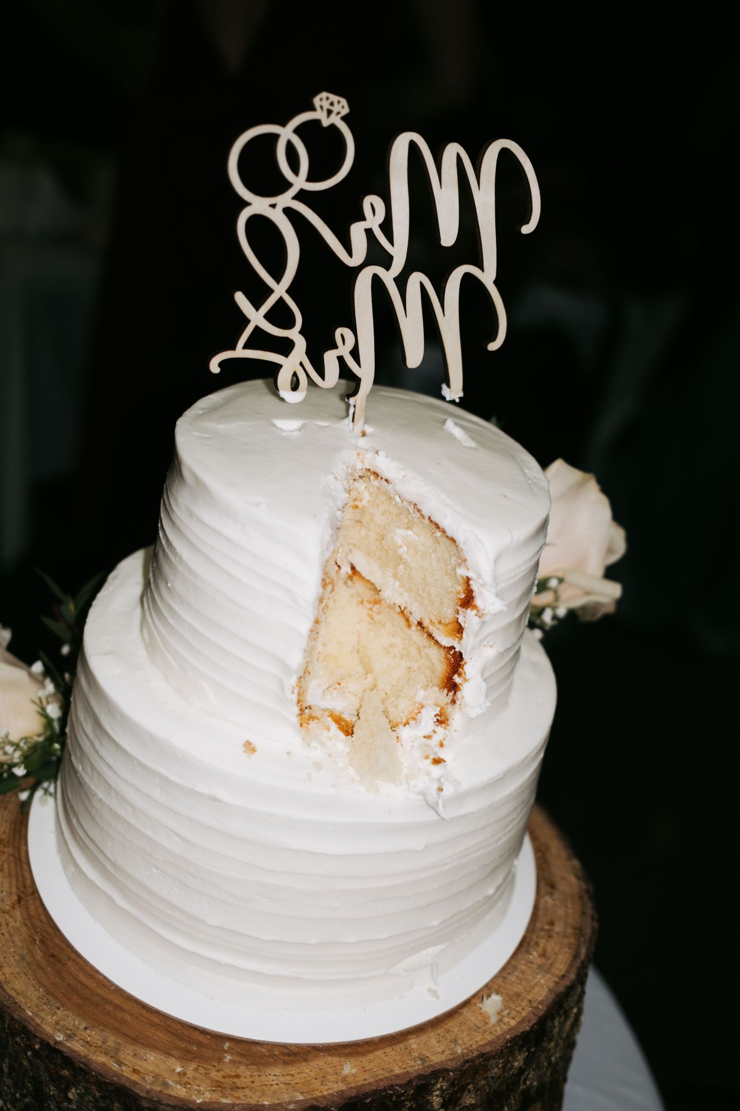 White wedding cake topper with decorative text displayed on a rustic wooden cake stand in progressive stages of assembly.