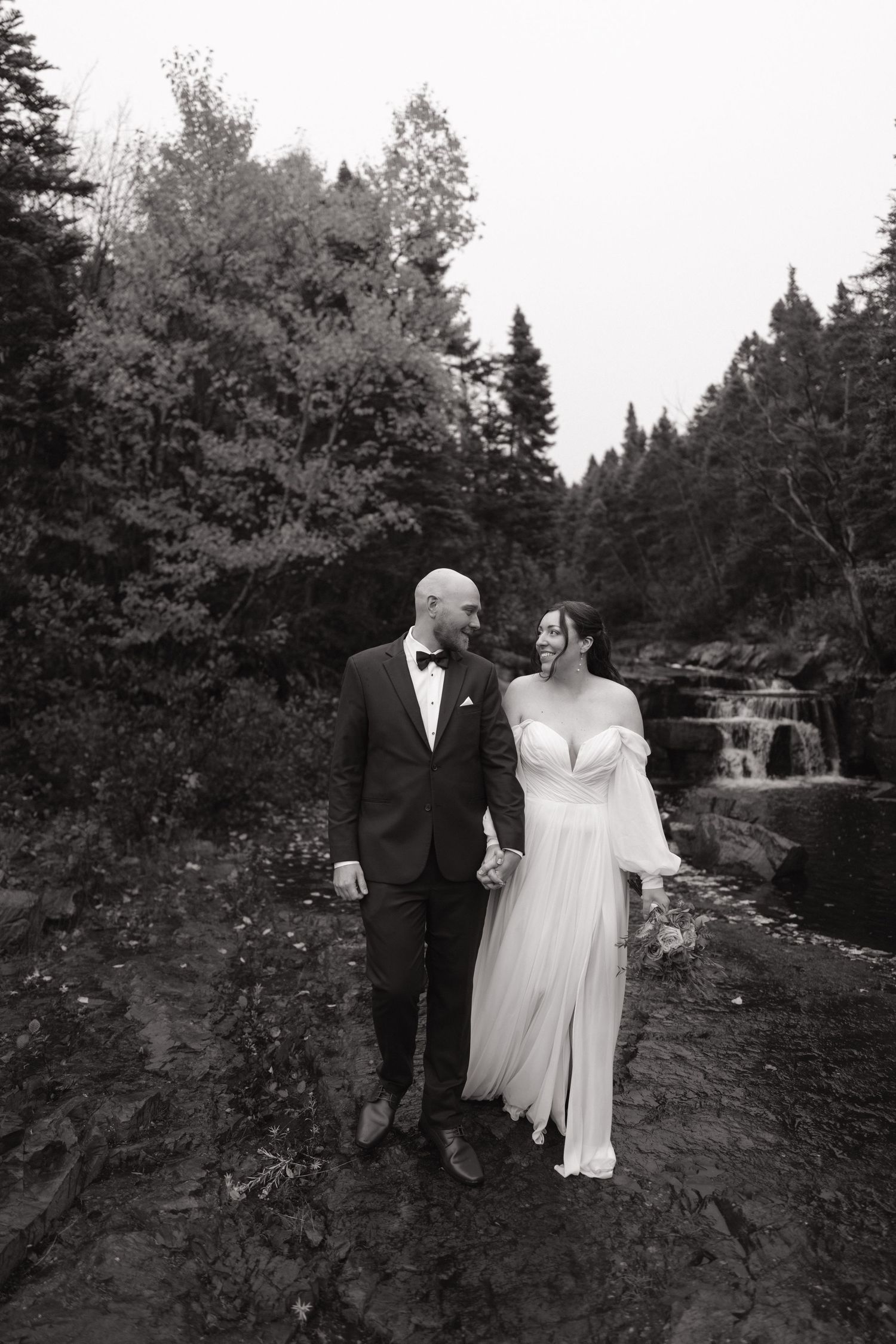 A couple in wedding attire walks together through a scenic wooded path surrounded by evergreen trees.