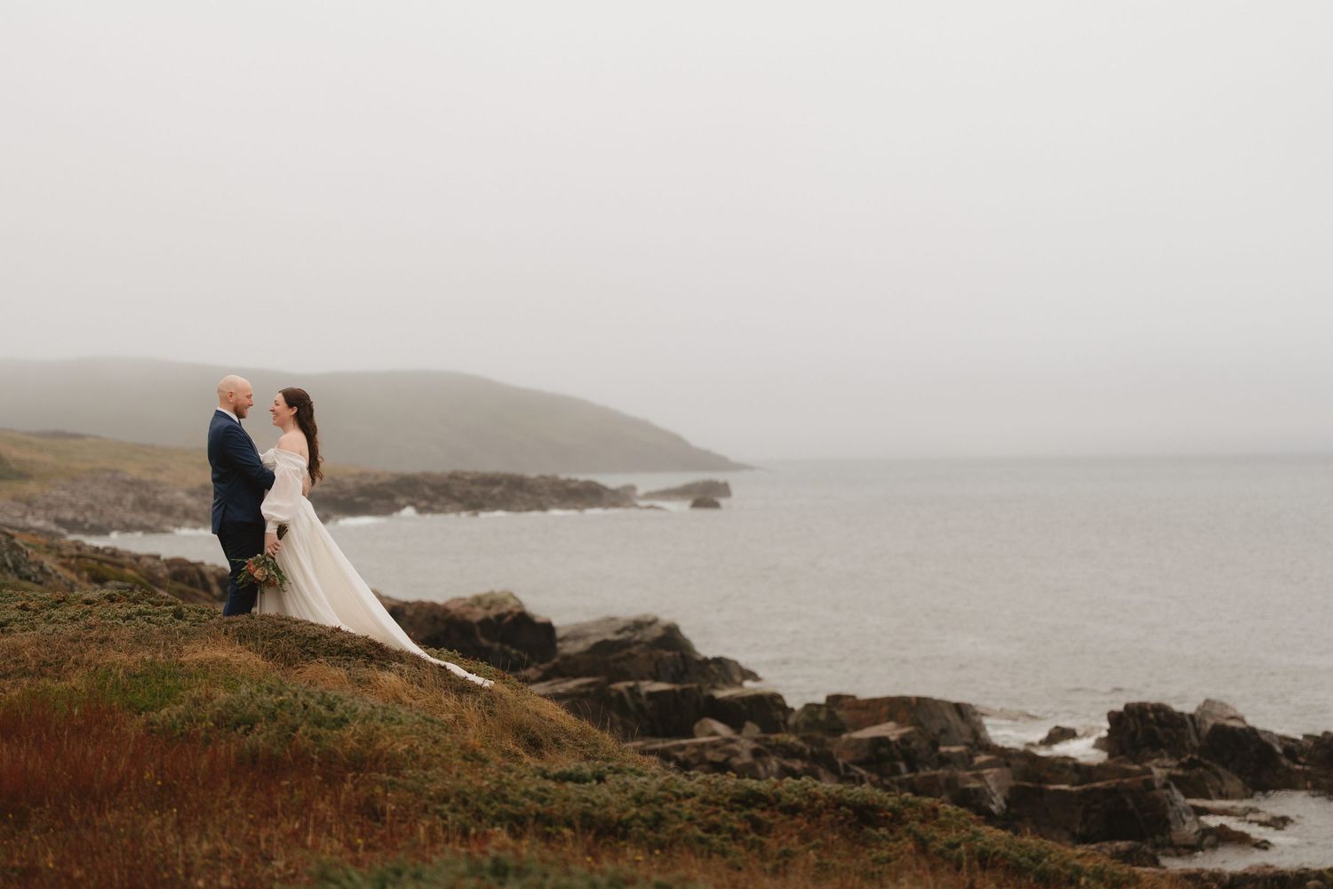 Couple in wedding attire embraces on a misty cliff overlooking the dramatic coastal landscape and ocean below.