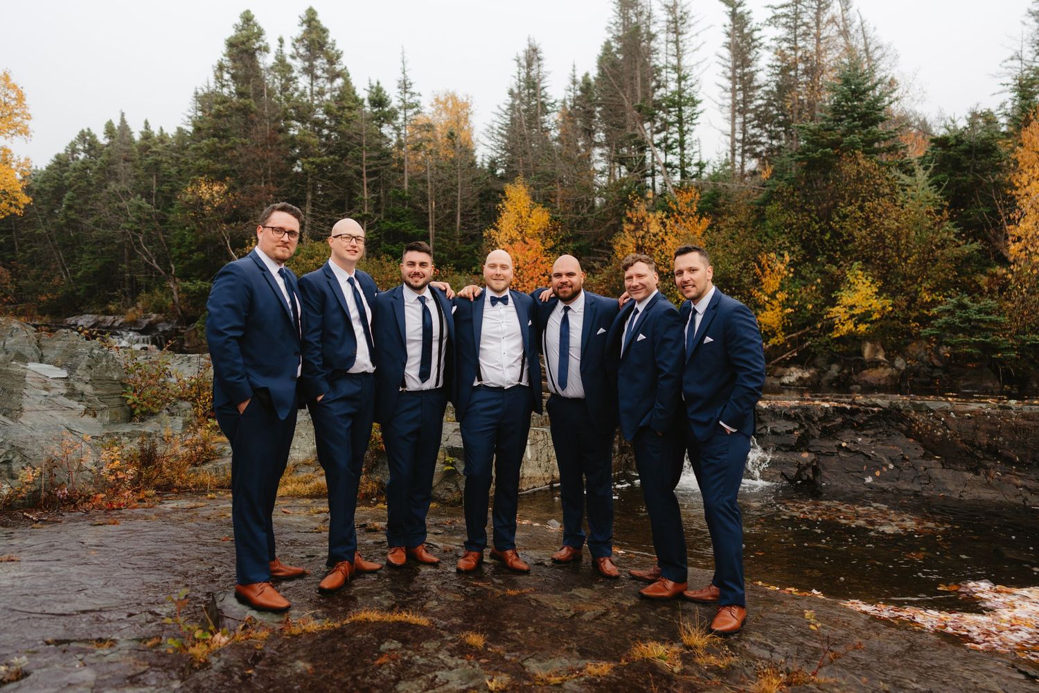 A group of groomsmen in navy suits stand together on rocky terrain with autumn foliage and evergreen trees in the background.