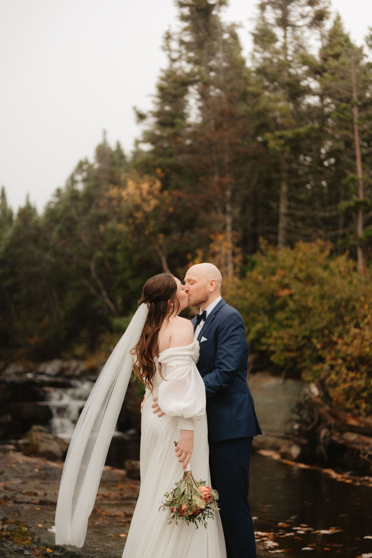 A romantic wedding couple shares a tender moment by a serene lake surrounded by evergreen trees in autumn.