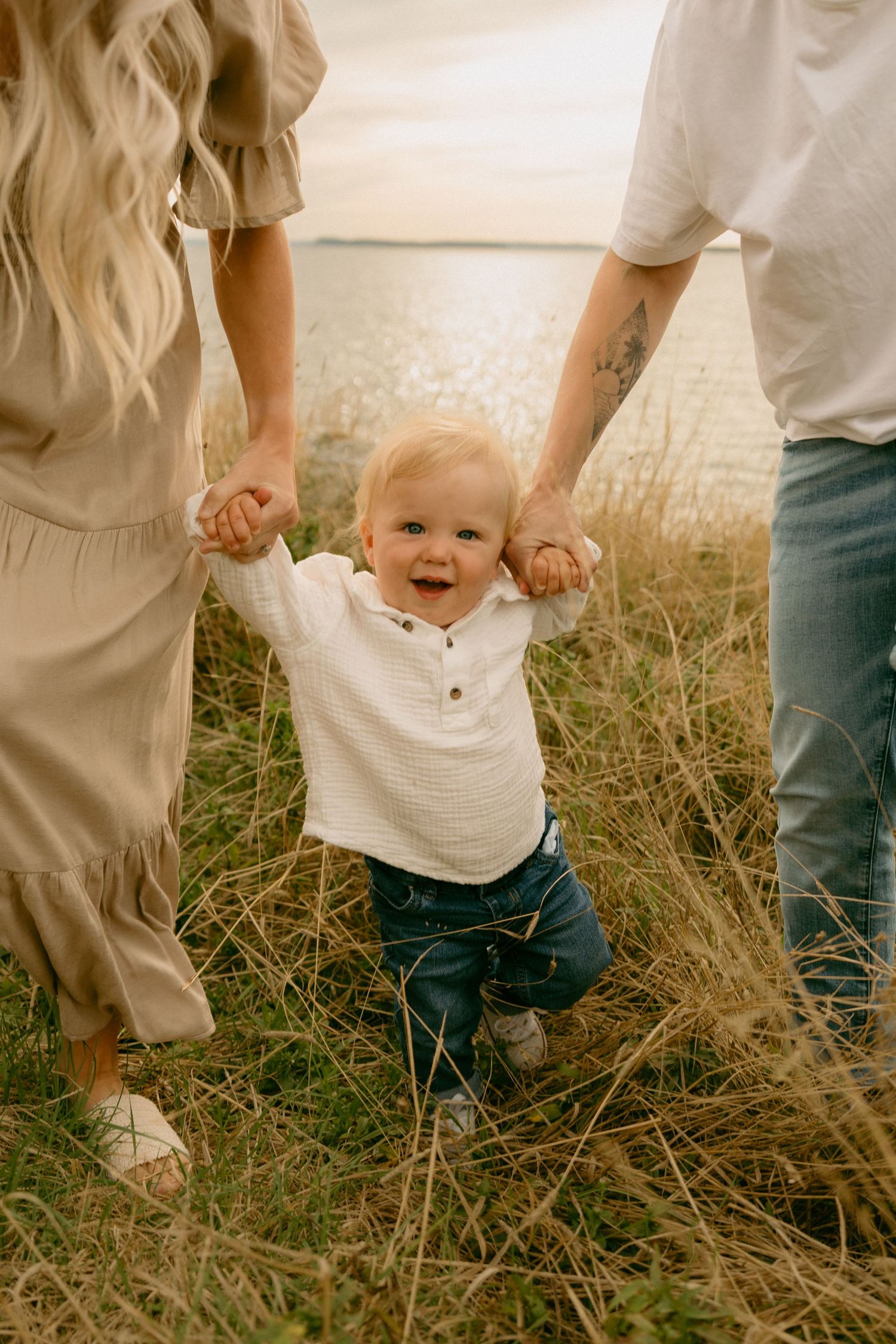 A small child learning to walk is held by supportive hands while standing in tall grass during golden hour.