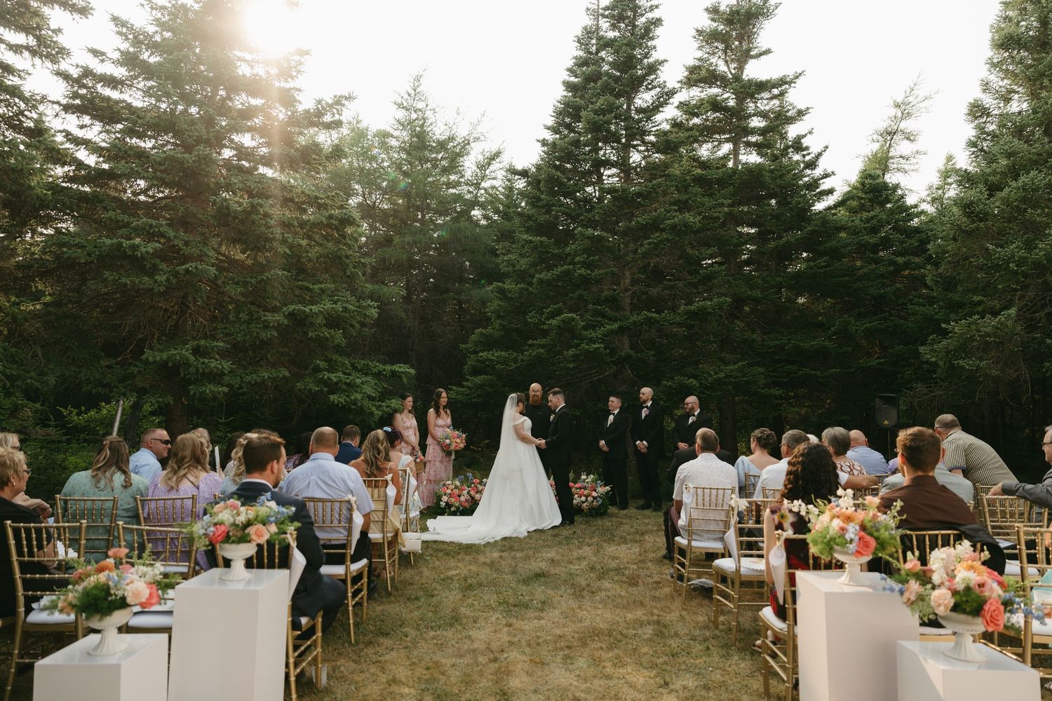 Scenic outdoor wedding ceremony site with guests seated among evergreen trees at sunset.
