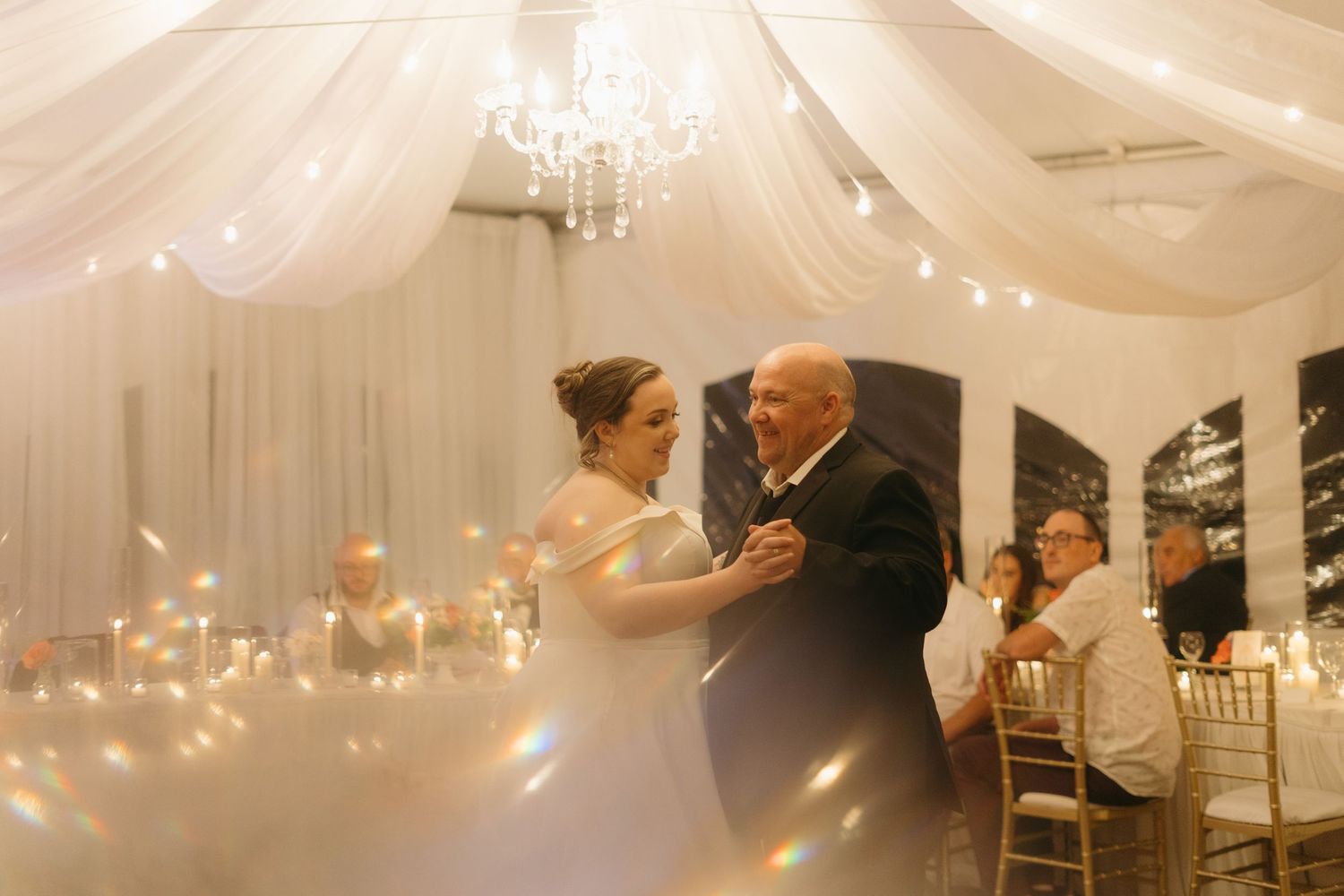 Couples dancing at an elegant wedding reception under white fabric draping and crystal chandeliers.