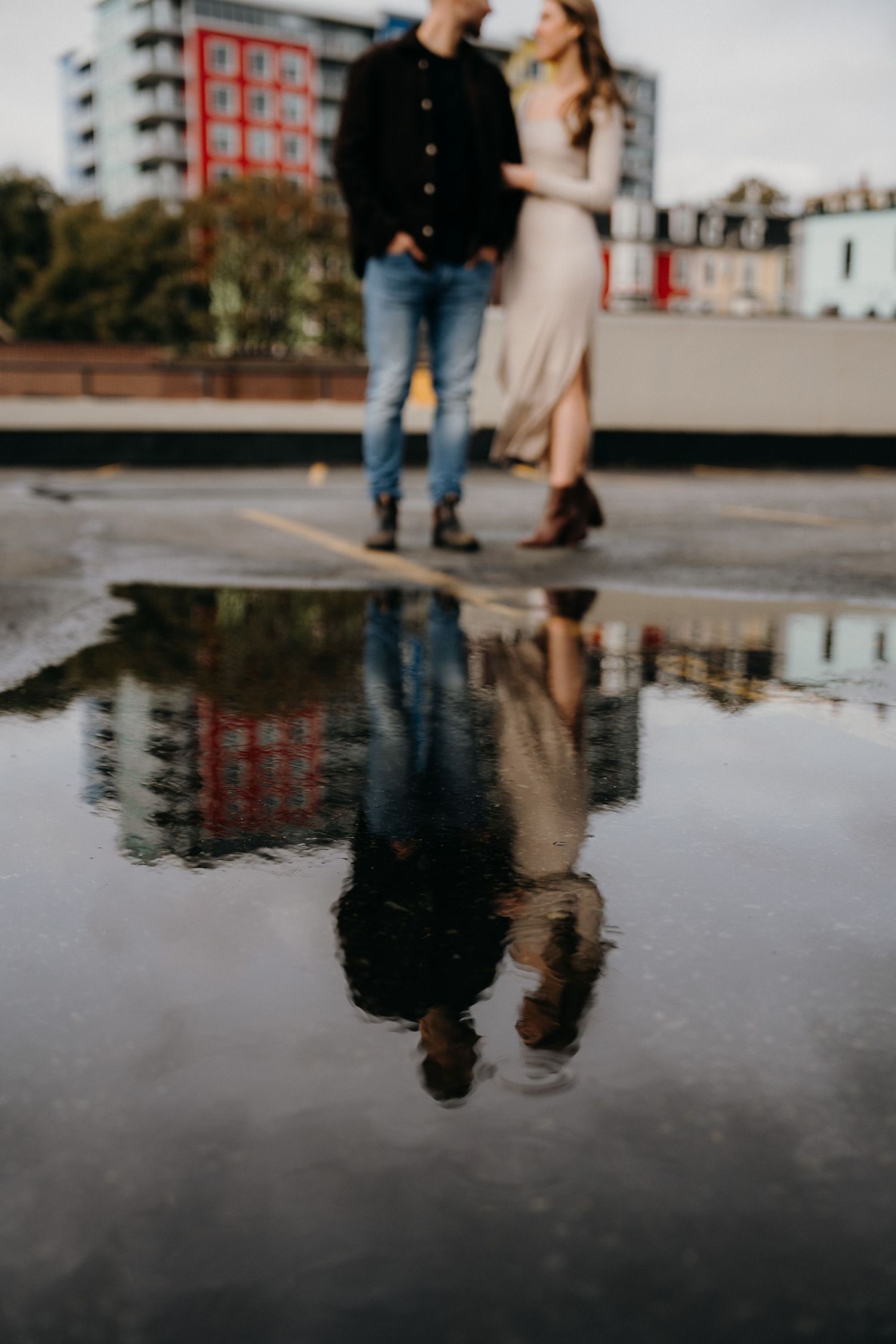 Intimate rooftop portrait with reflection in standing water and red building in background.