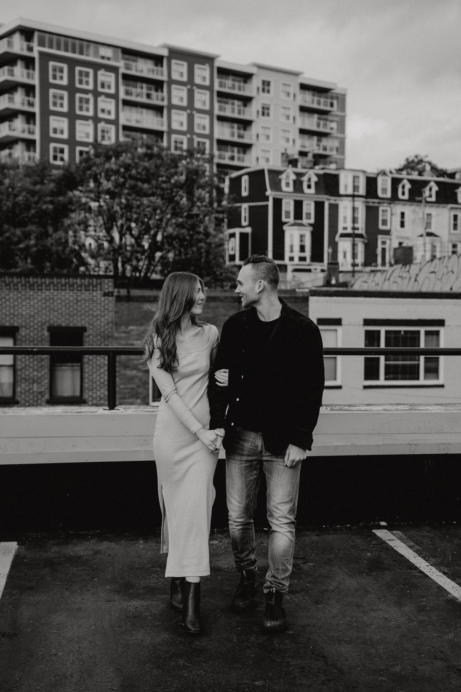 Black and white photo of couple walking together on urban rooftop with apartment buildings visible.
