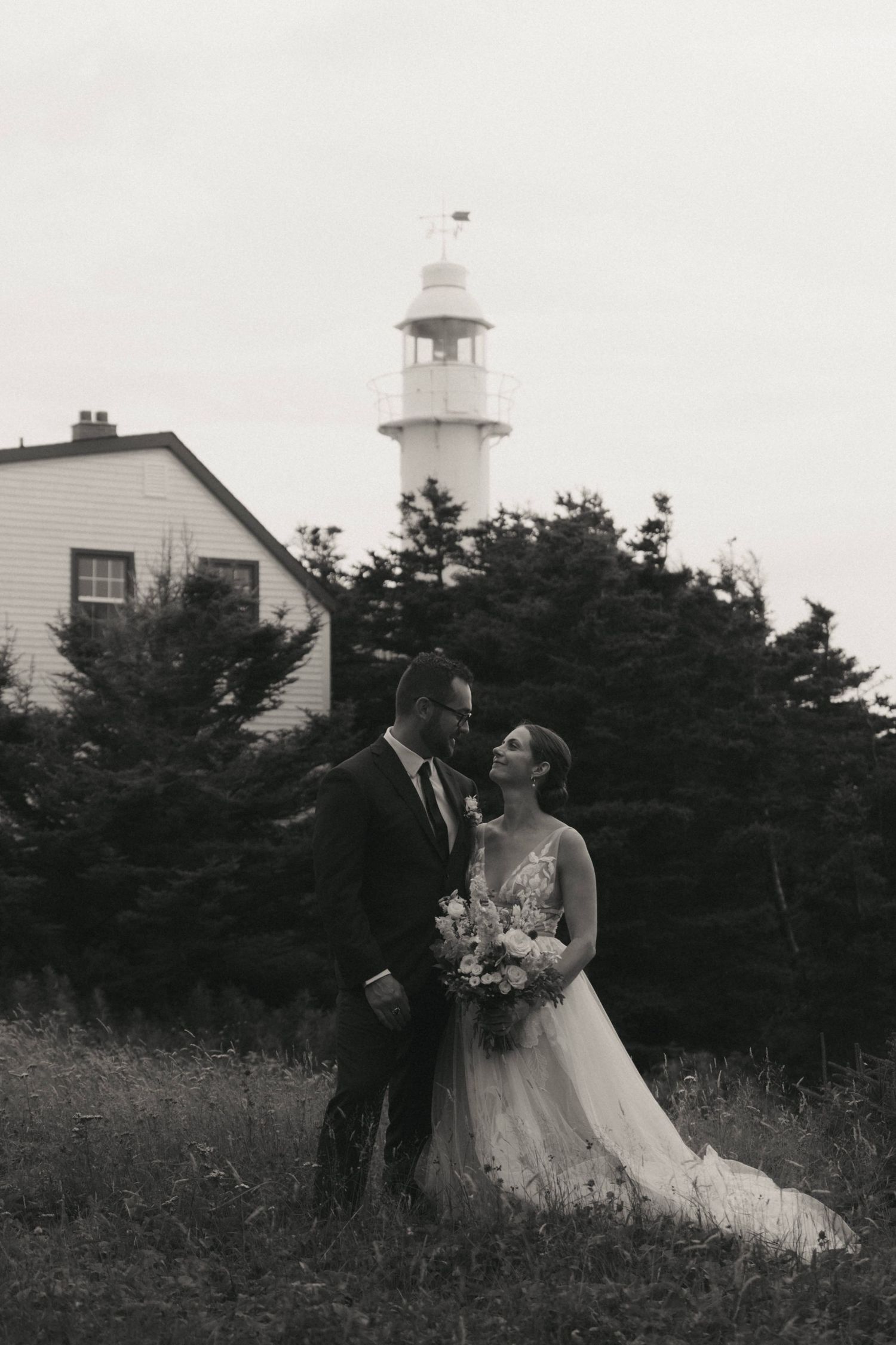 Black and white wedding couple poses near a lighthouse in a field of tall grass and wildflowers at dusk.