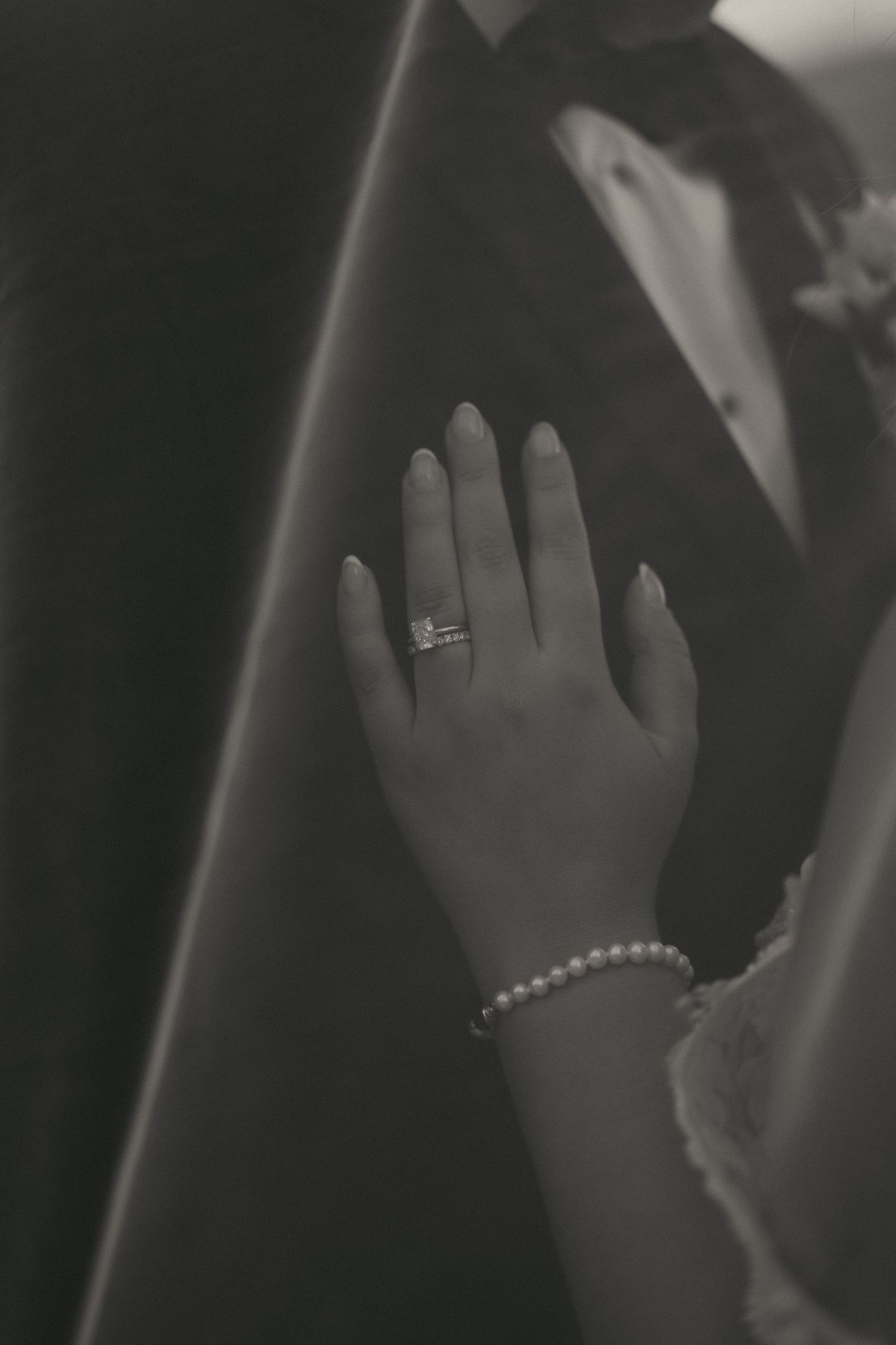 Black and white photo of a womans hand with engagement ring and pearl bracelet resting on a mans suit jacket.
