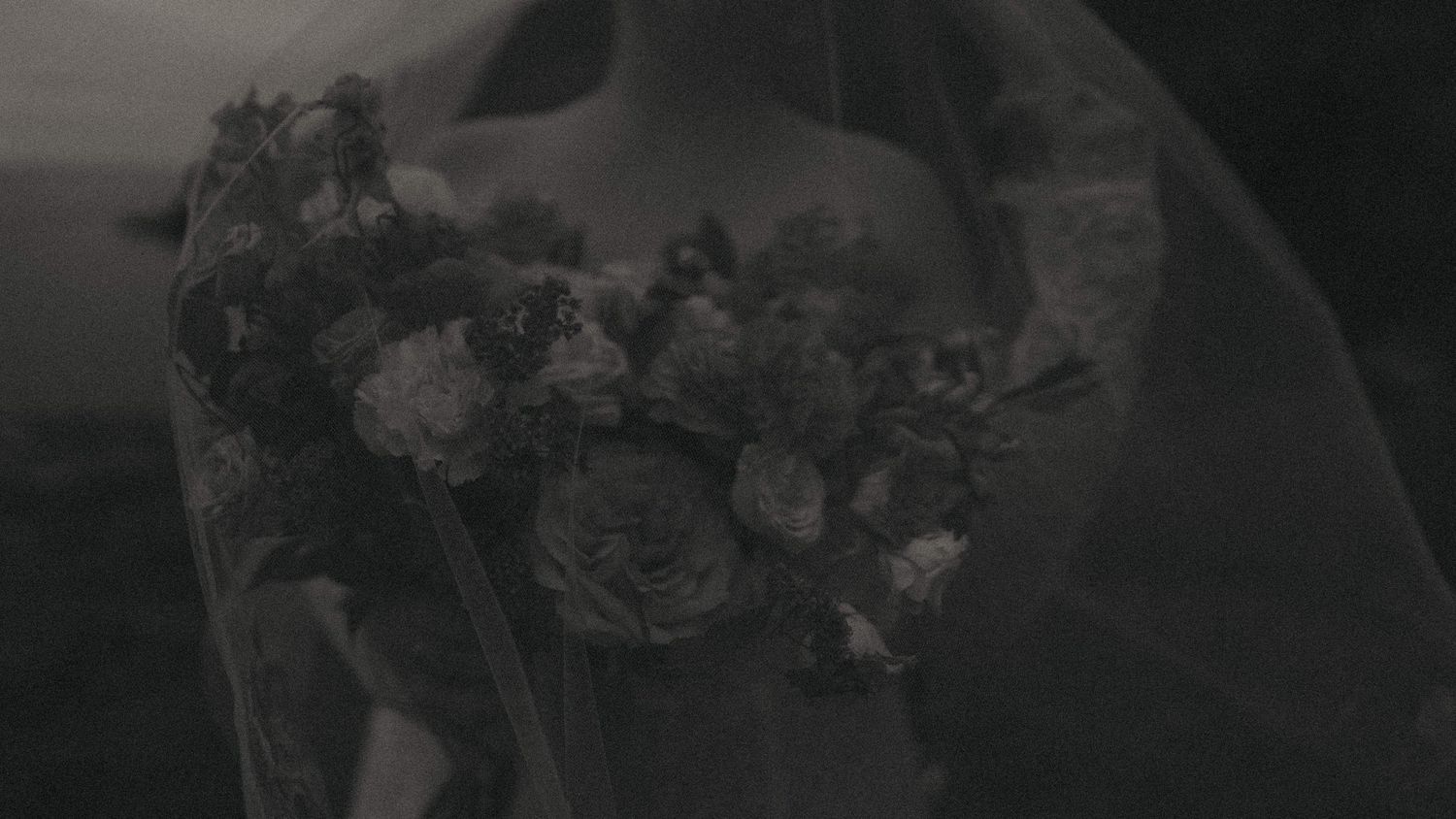 Bride holding a lush floral bouquet with roses under a veil in a moody black and white wedding photo.