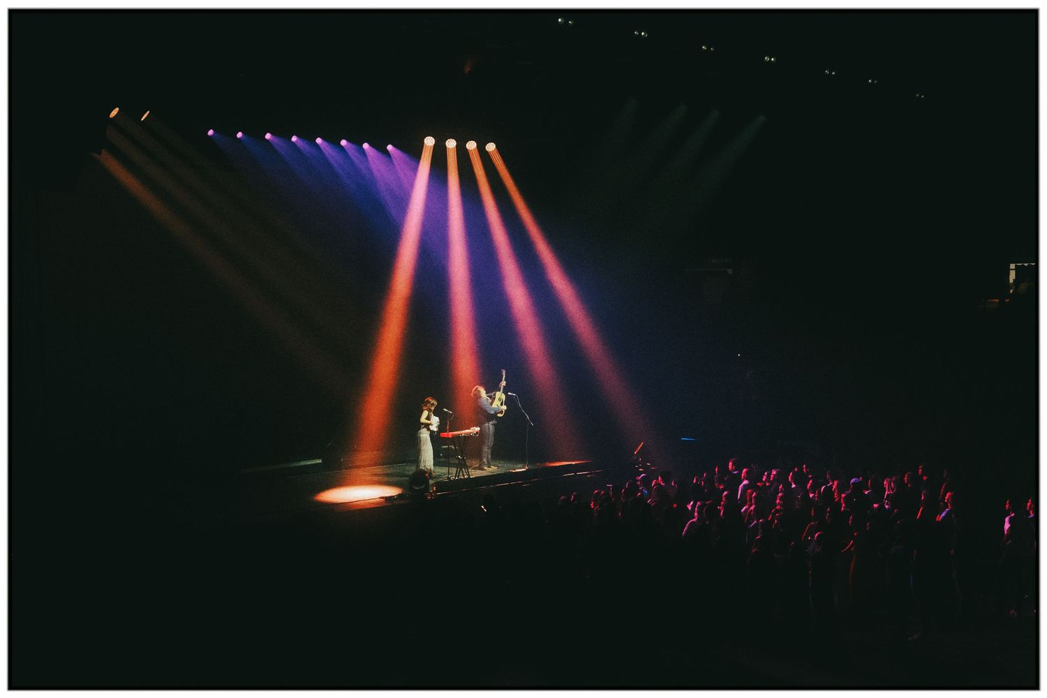 Concert stage illuminated with dramatic purple and red spotlights during a live performance with silhouetted figures.