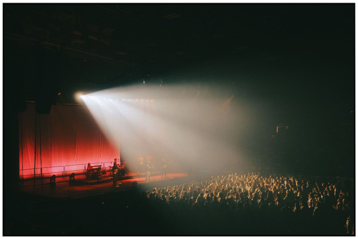 A bright spotlight beam illuminates a dark concert venue with a red-lit stage and packed crowd of silhouetted spectators.