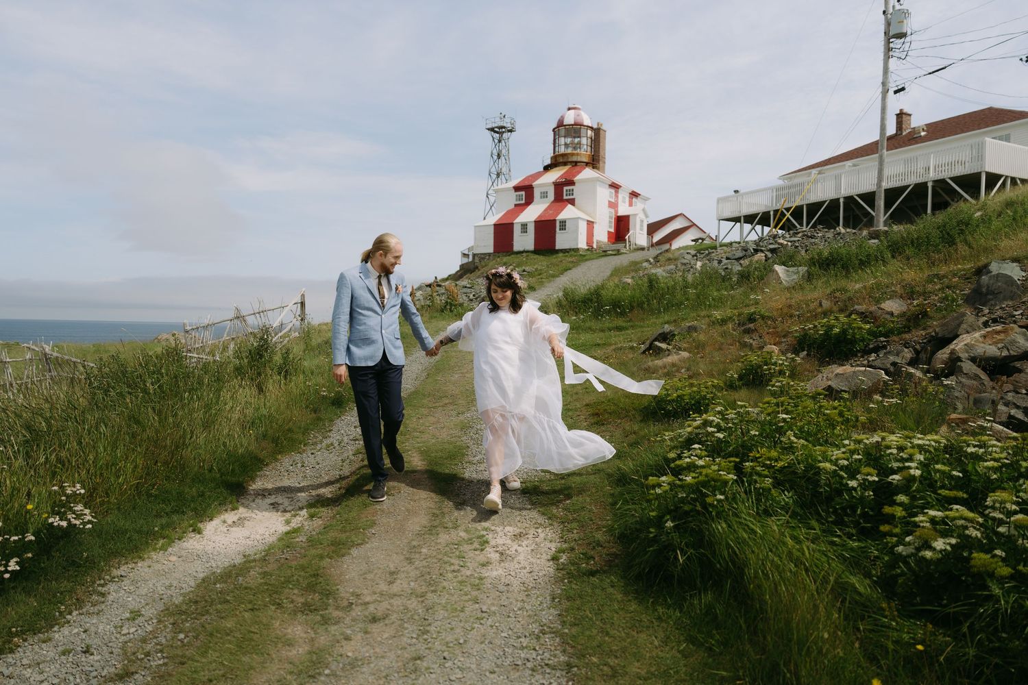 Couple in wedding attire walks along scenic coastal path with lighthouse and ocean views in background.