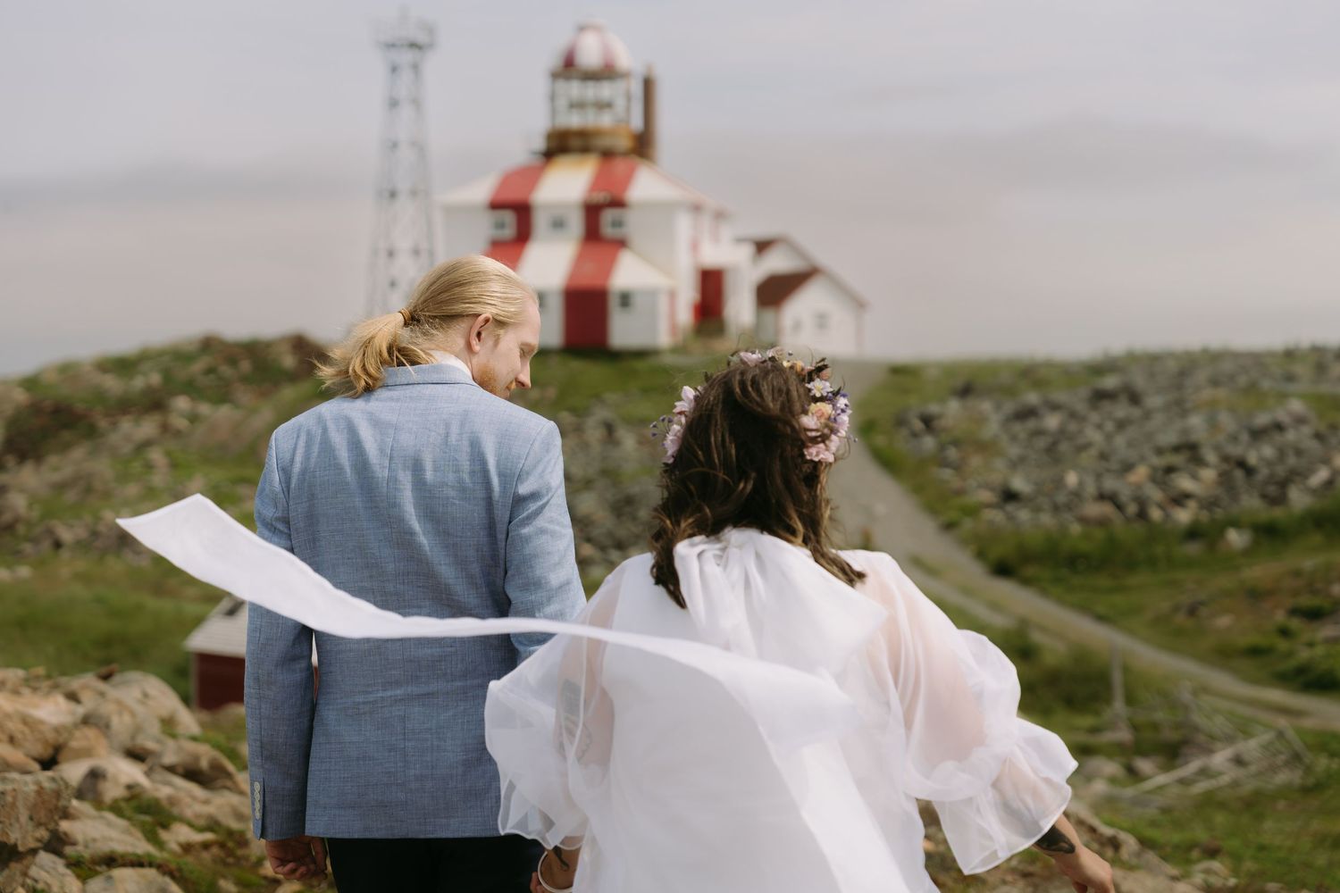 A couple in wedding attire stands on a coastal cliff overlooking a lighthouse and ocean view.