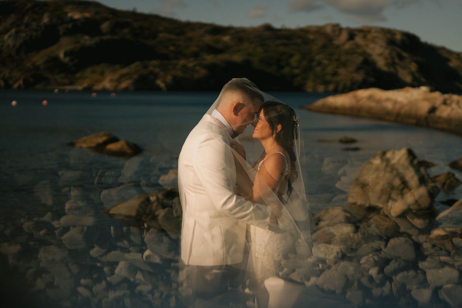 Romantic couple embraces on rocky beach at sunset with dramatic coastal cliffs in background.