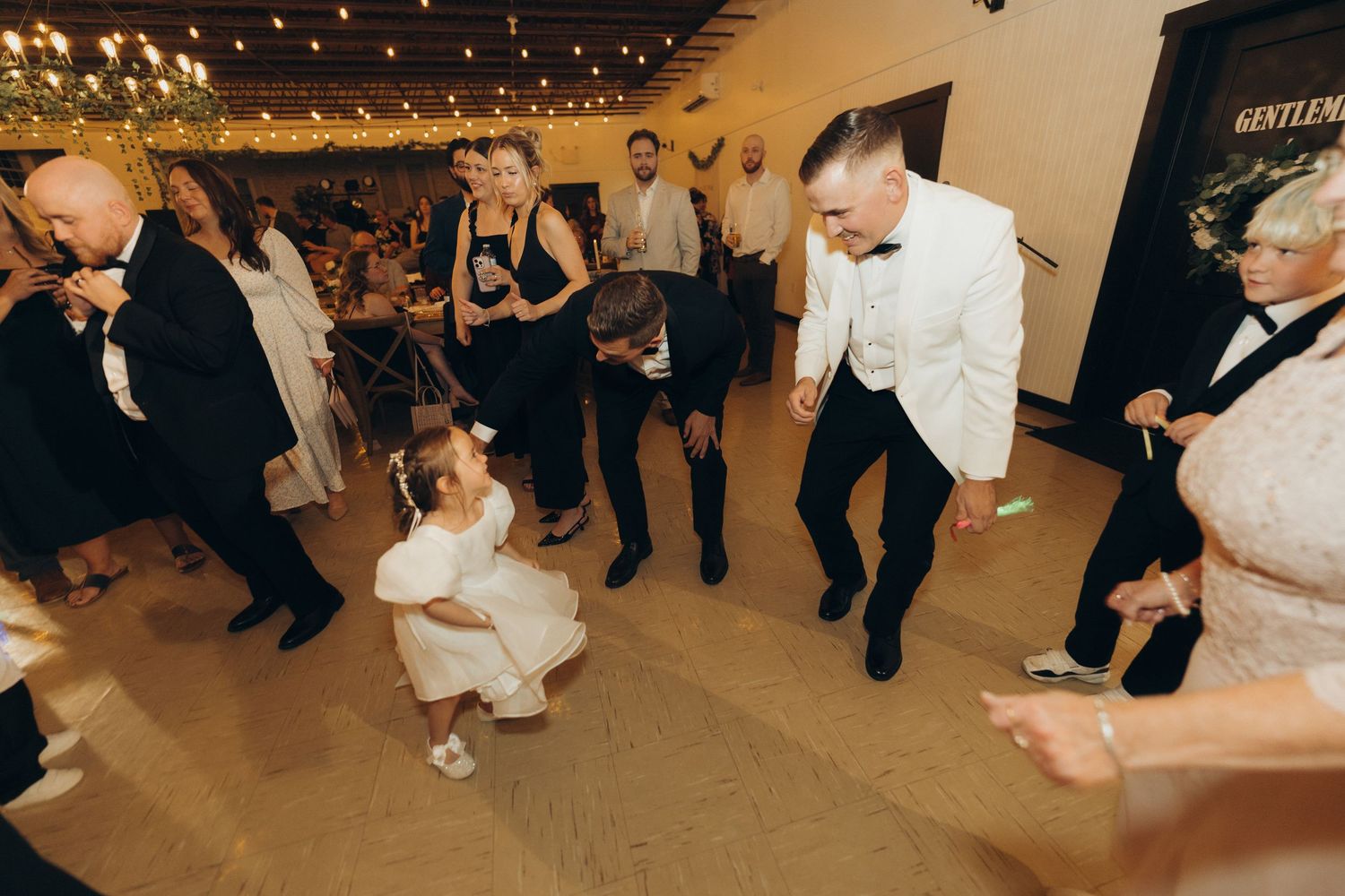 Wedding guests dance and celebrate on a rustic wooden floor under warm string lights at an indoor reception.