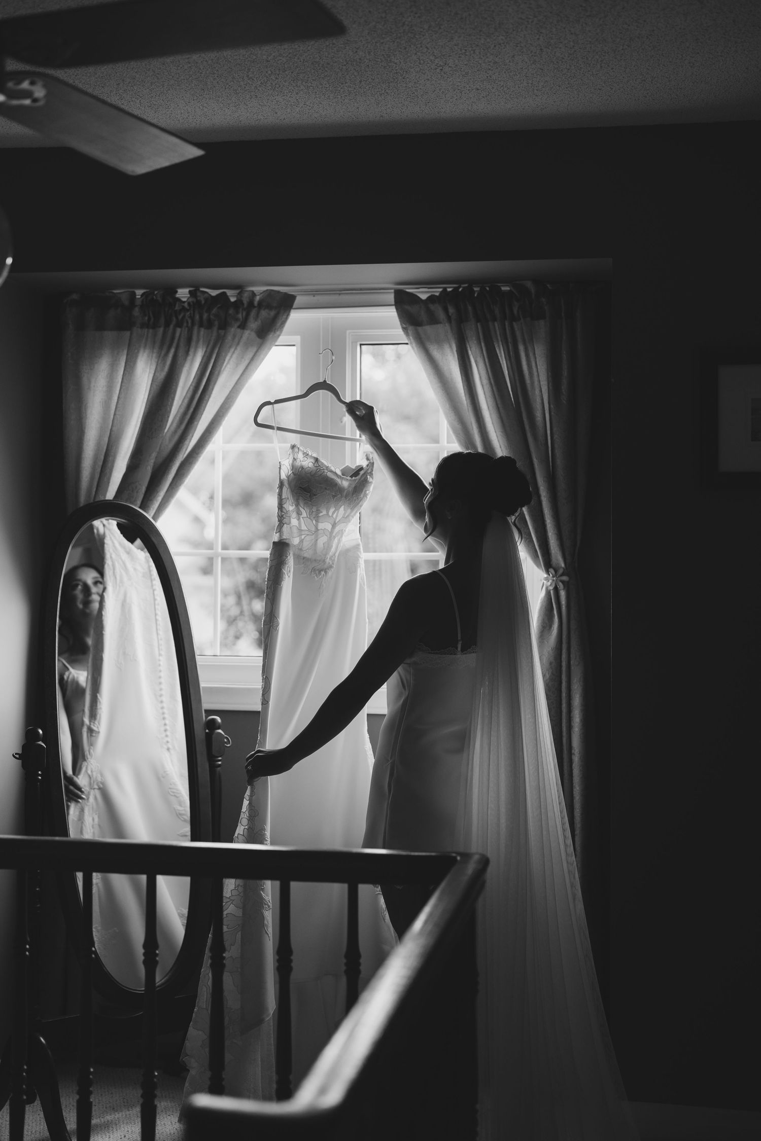 Black and white photo of a bride holding her wedding dress on a hanger near a window, her reflection visible in a mirror.