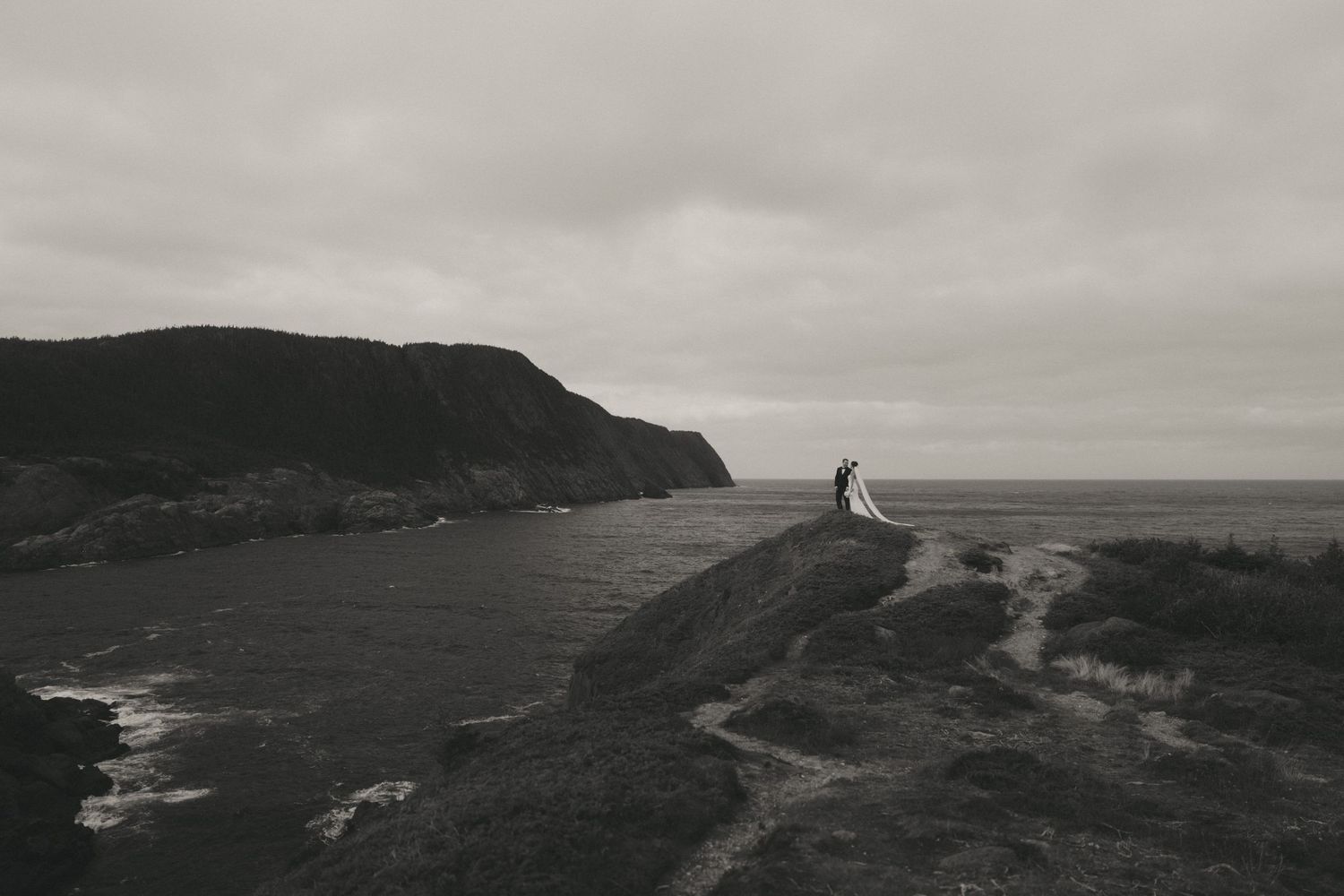 Couple in wedding attire standing on rocky coastal cliffs with dramatic overcast skies and ocean views in black and white.