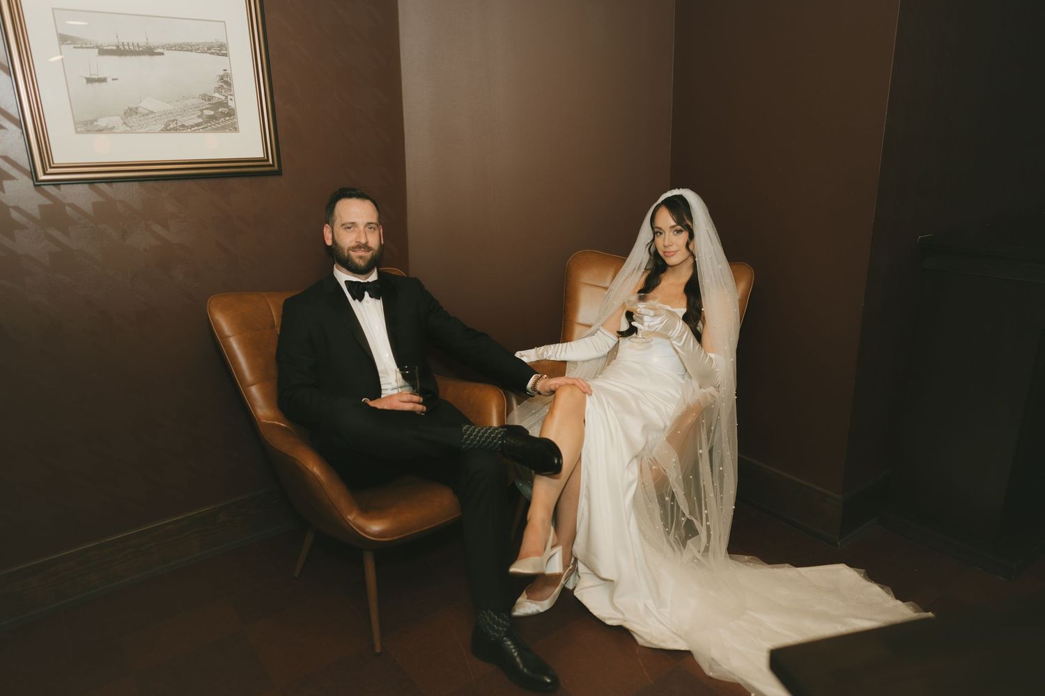 Bride and groom sitting in leather chairs wearing wedding attire, white gown with veil and black tuxedo.