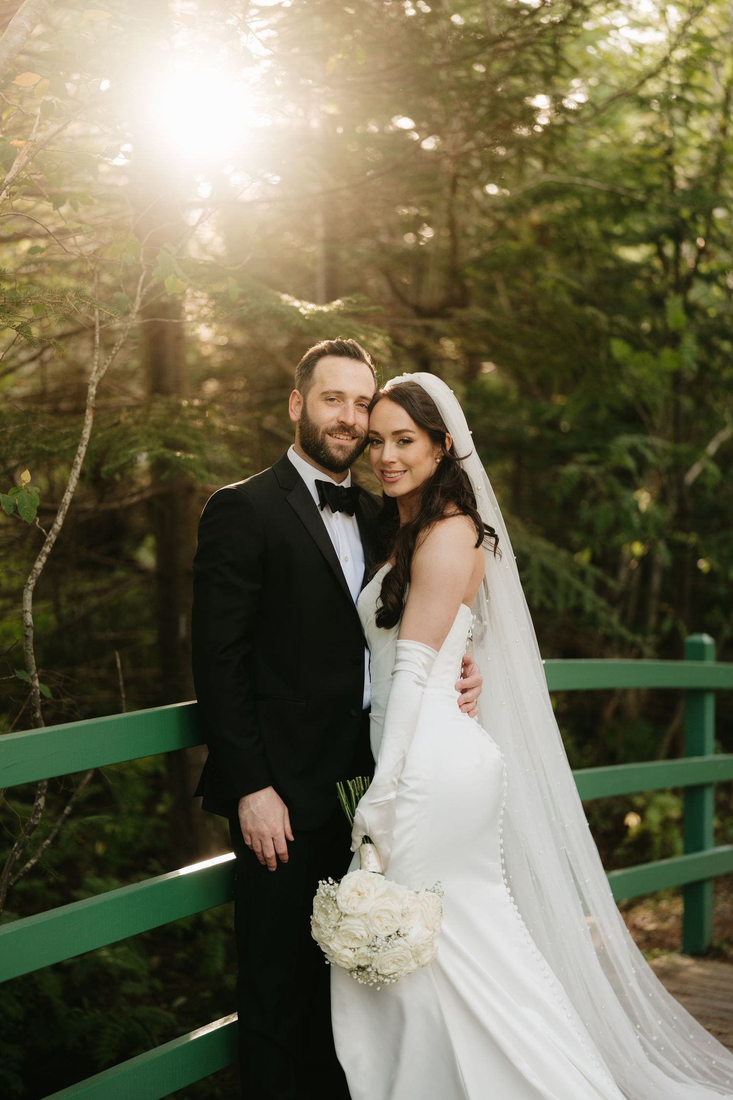 Bride and groom pose on a green bridge surrounded by sunlit forest, bride holding white bouquet in elegant gown with veil.