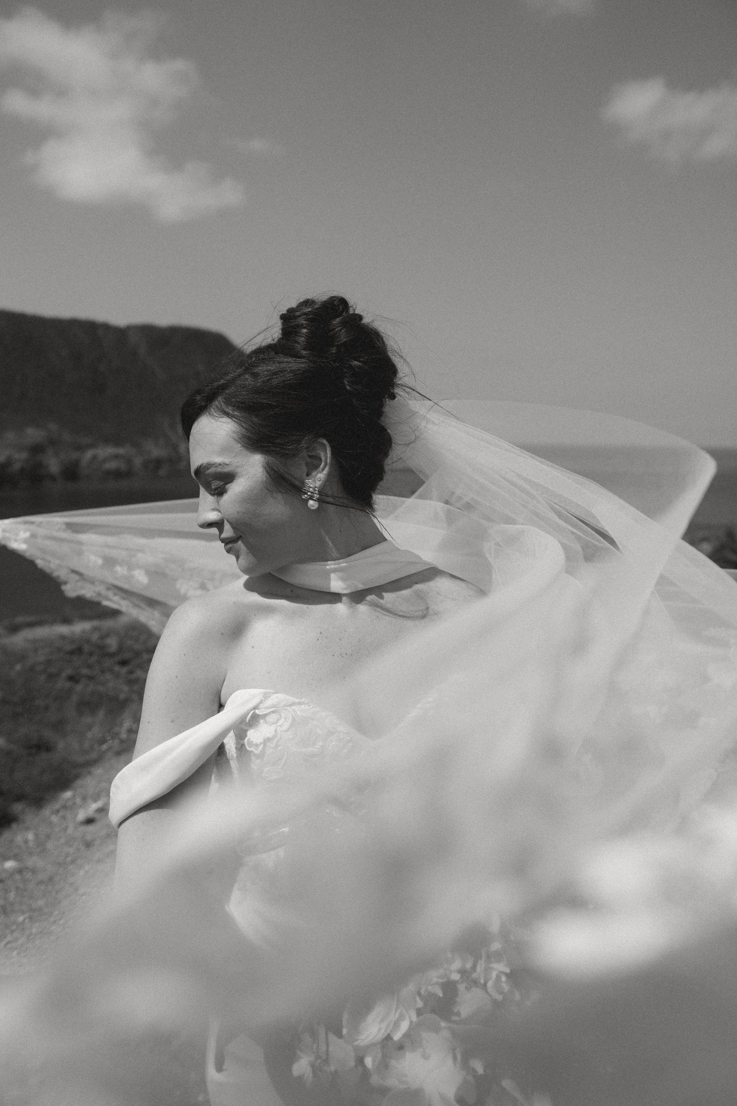 Black and white photo of a bride in a flowing veil and strapless gown, hair up, smiling outdoors on a windy day.