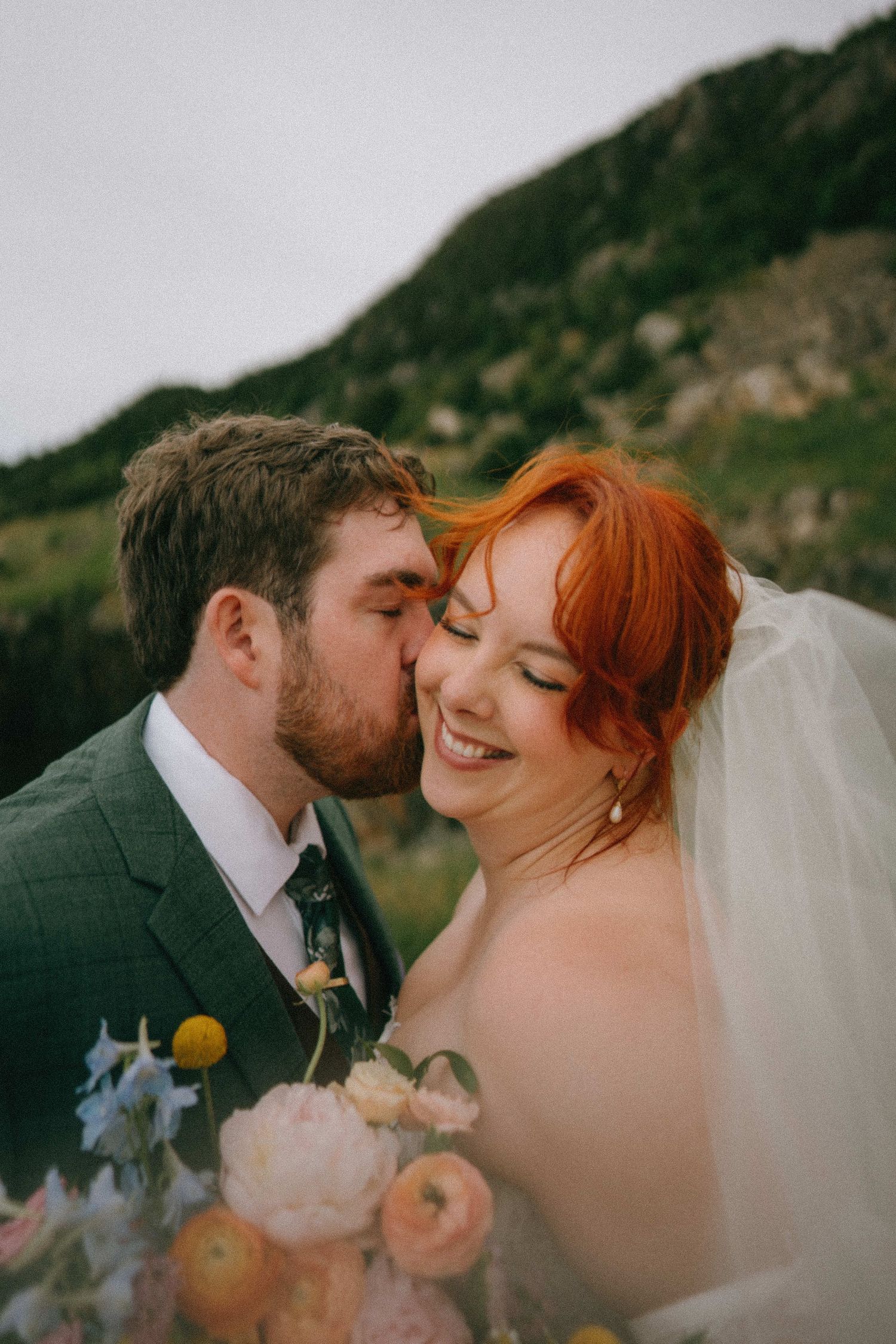 Groom in green suit kisses bride with red hair wearing veil, holding colorful bouquet with mountain backdrop.