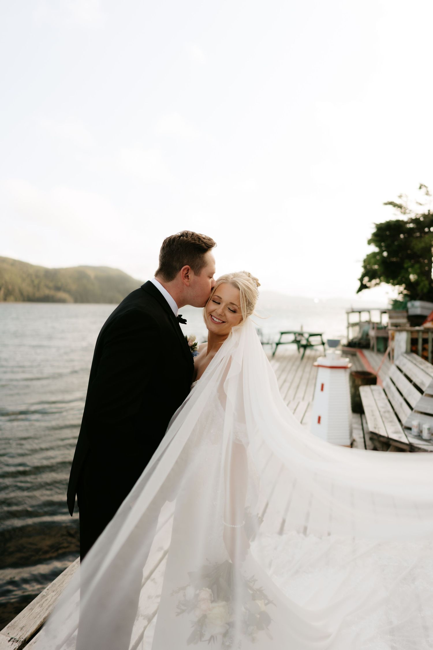Bride and groom share a romantic moment on a lakeside dock, her veil flowing in the breeze, with scenic hills behind.
