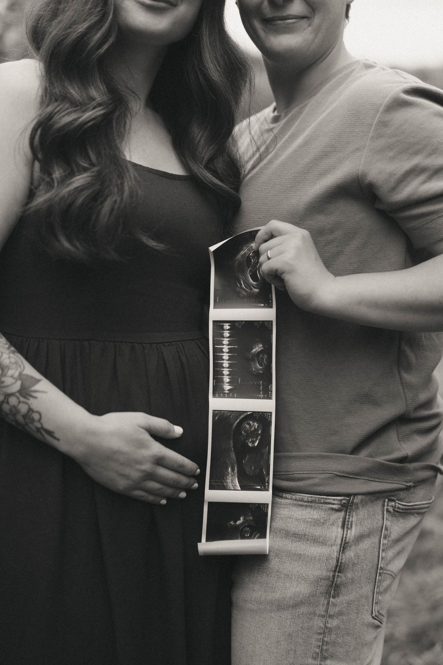 Black and white photo of pregnant couple holding ultrasound images outdoors, woman in black dress with tattoo.