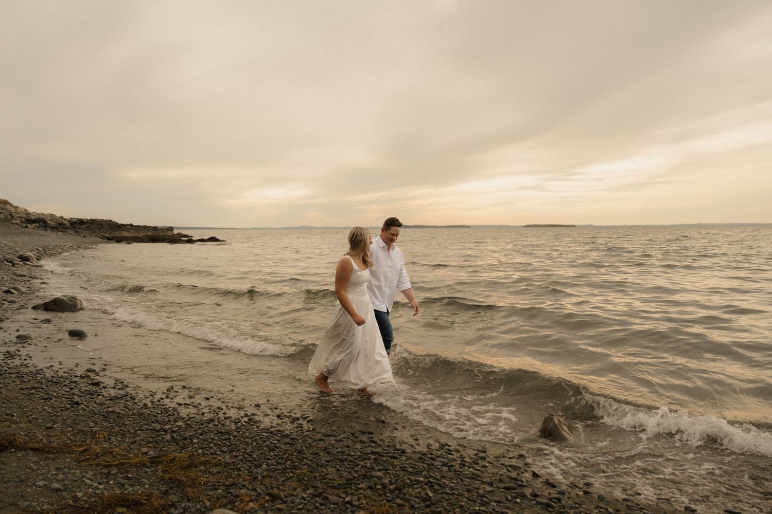 Couple walking along rocky beach shoreline during golden sunset with waves crashing against the coast.