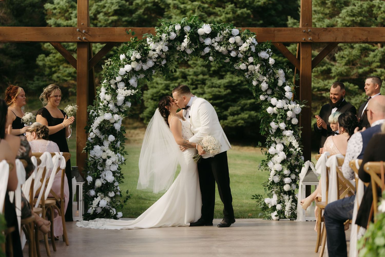 A wedding ceremony takes place under a rustic wooden pergola decorated with white floral arrangements in an outdoor garden setting.