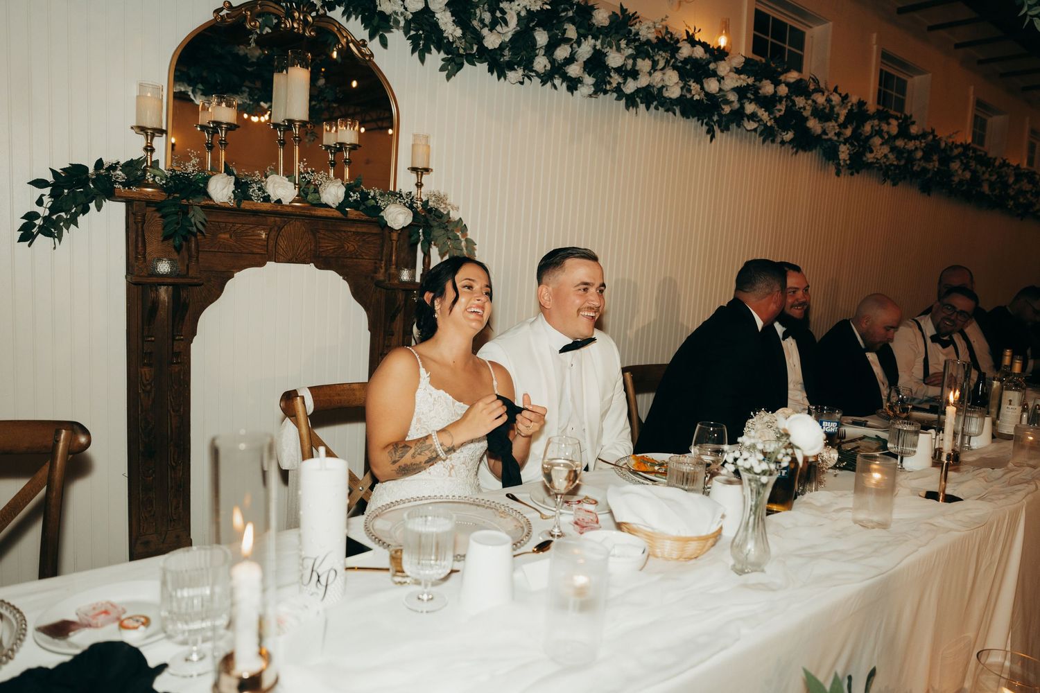 Wedding reception guests seated at long tables decorated with greenery and white linens in an elegant indoor venue.