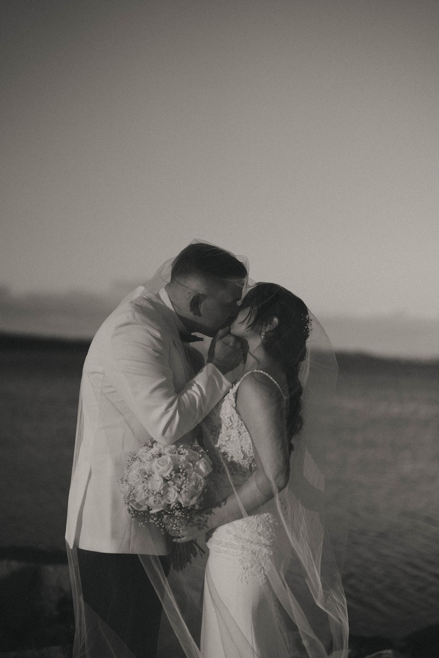 A romantic black and white photo series showing a couple in wedding attire embracing by the ocean at sunset.