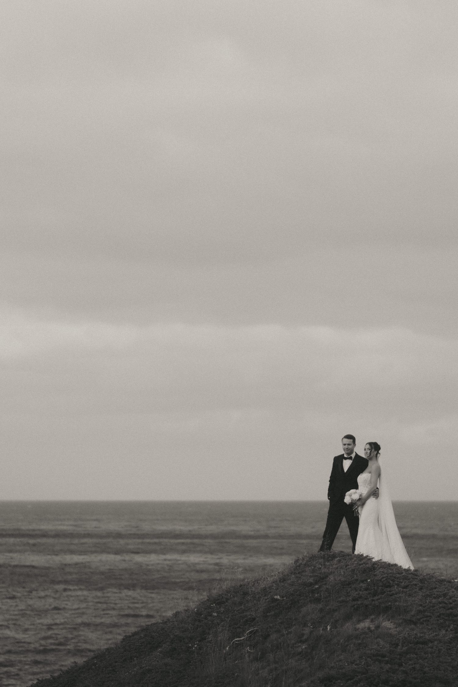 Black and white photo of bride and groom standing on coastal cliff edge with dramatic overcast sky and ocean backdrop.