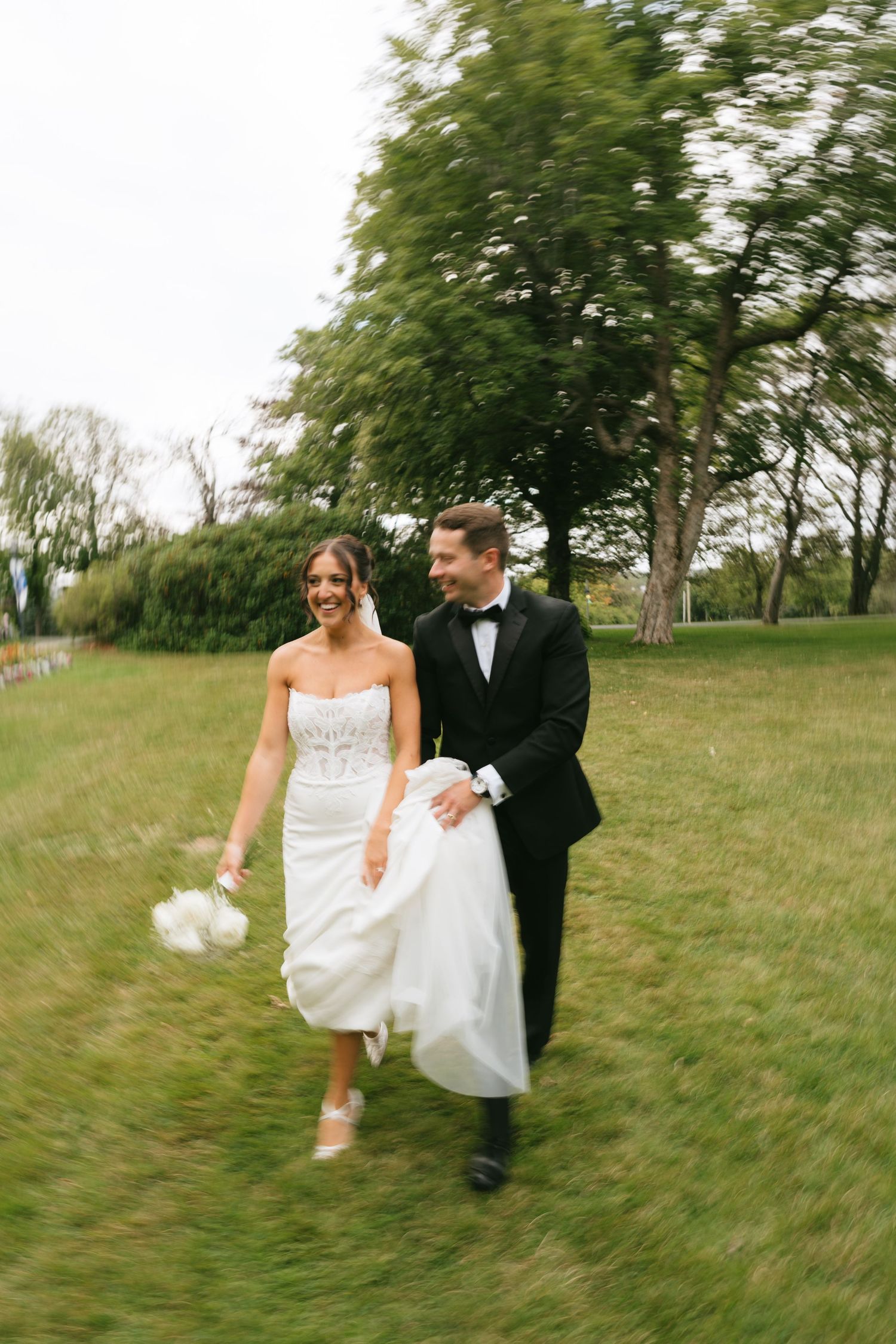 A smiling bride in a strapless white gown and groom in black tuxedo walk joyfully through a lush green park.