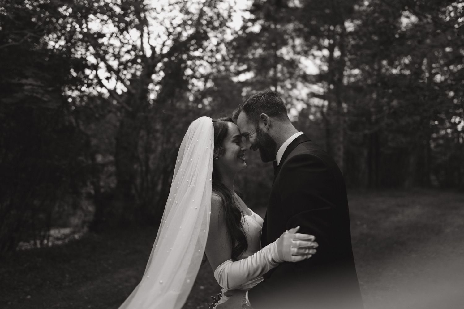 Black and white photo of a bride and groom sharing an intimate moment, touching foreheads outdoors amid lush trees.