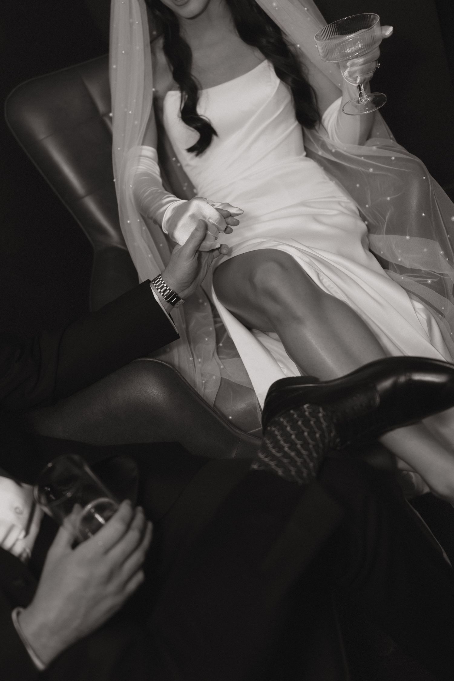 Black and white photo of bride in short white dress and veil holding champagne glass, seated beside groom in suit.