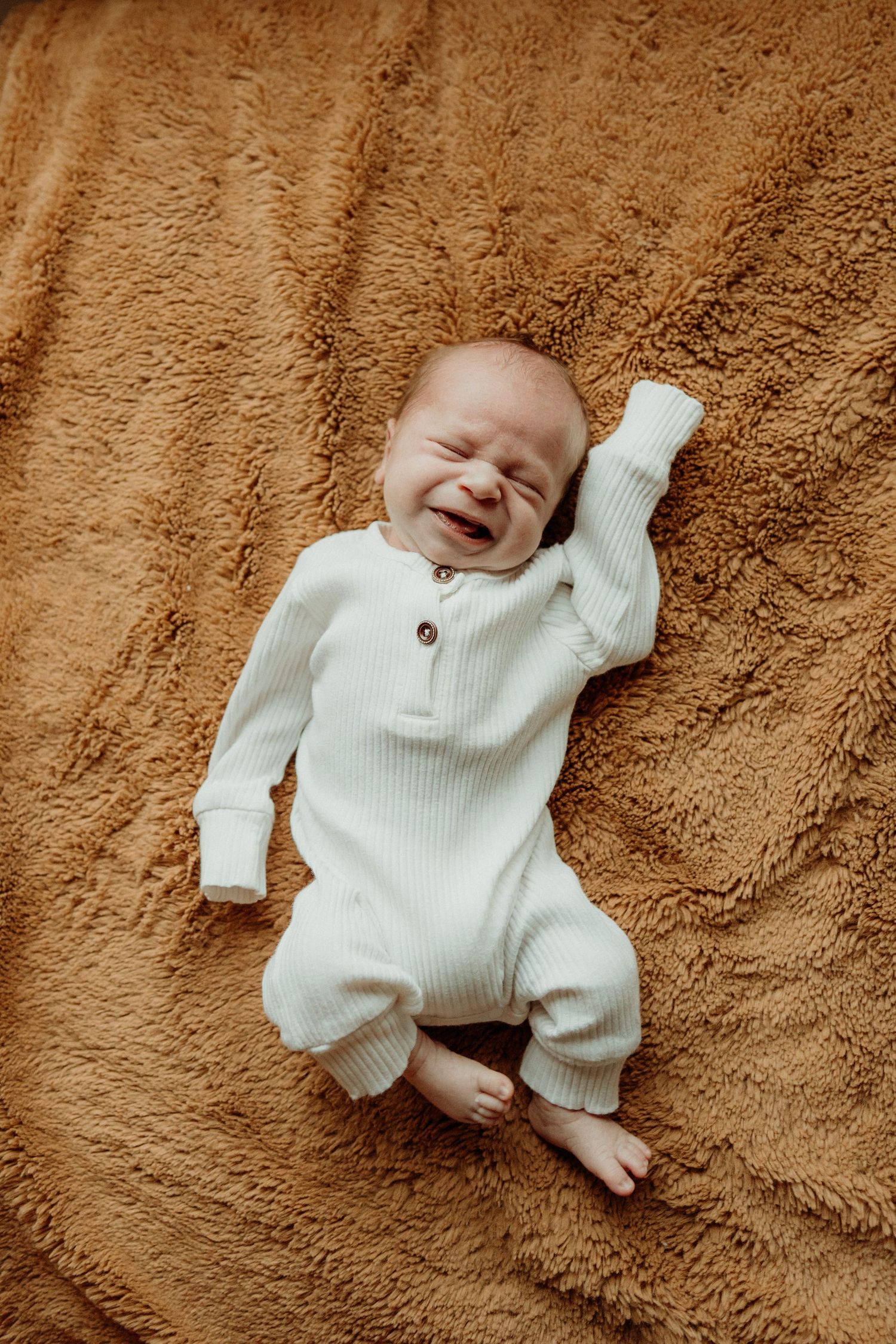 A baby in a white ribbed sweater lying on a soft brown blanket.