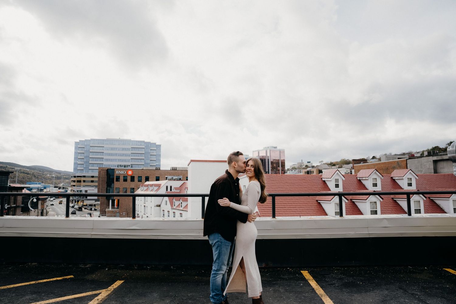 Couple embracing on an urban rooftop with industrial buildings and cloudy skies in the distance.