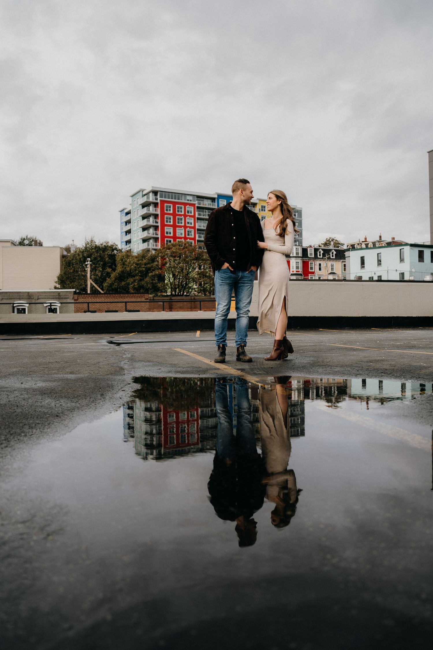 A couple poses by a reflective puddle on a rooftop with their mirror image in the water.