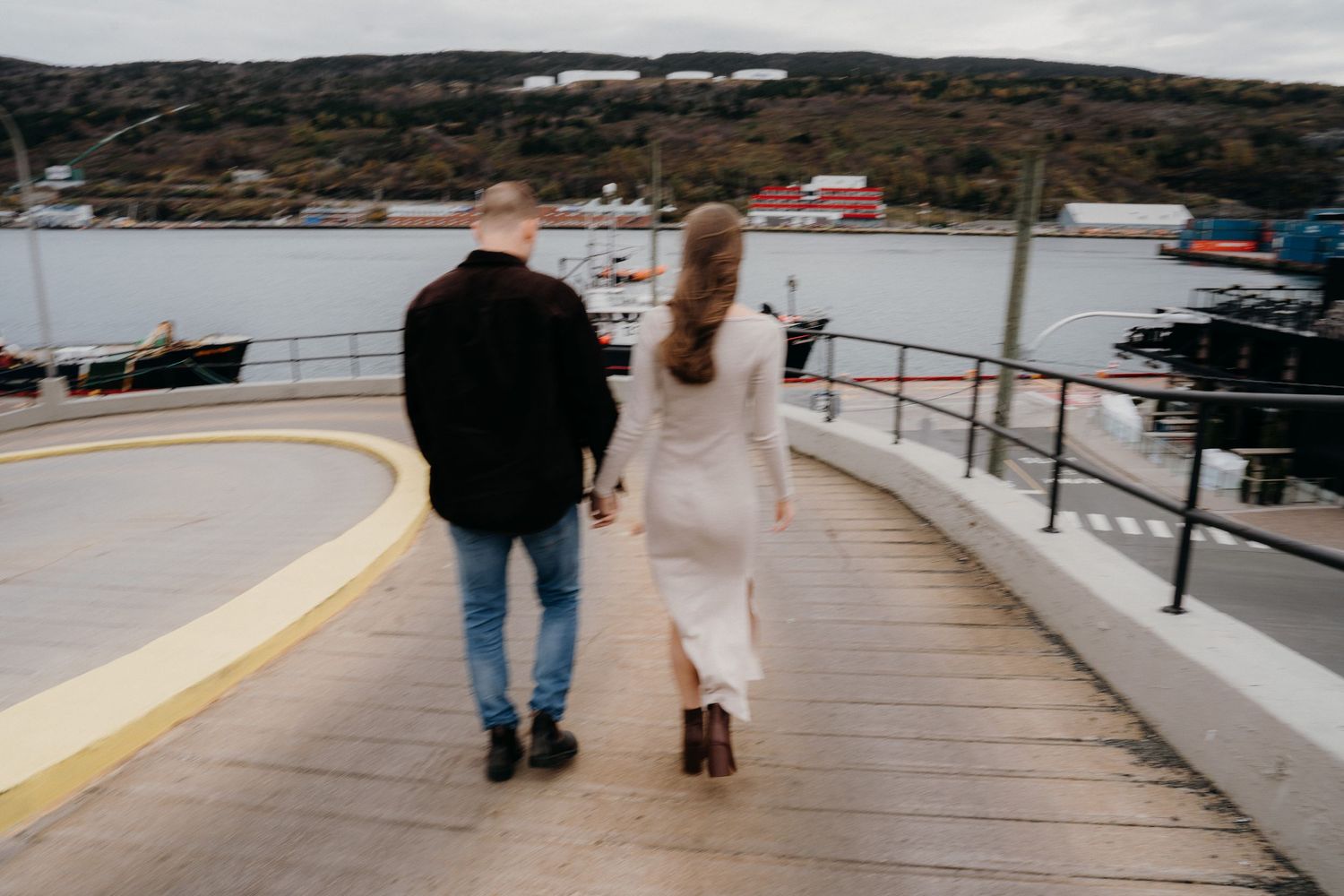 A couple walks along a curved waterfront boardwalk with mountains and buildings visible in the background.