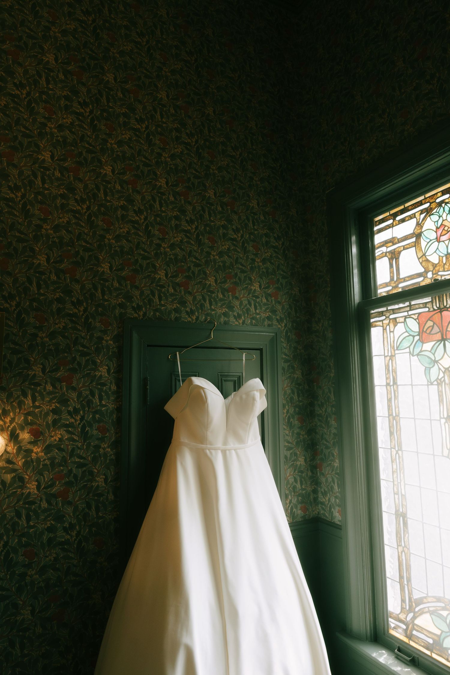 A white wedding dress hangs gracefully near a vintage window with decorative glass panes.