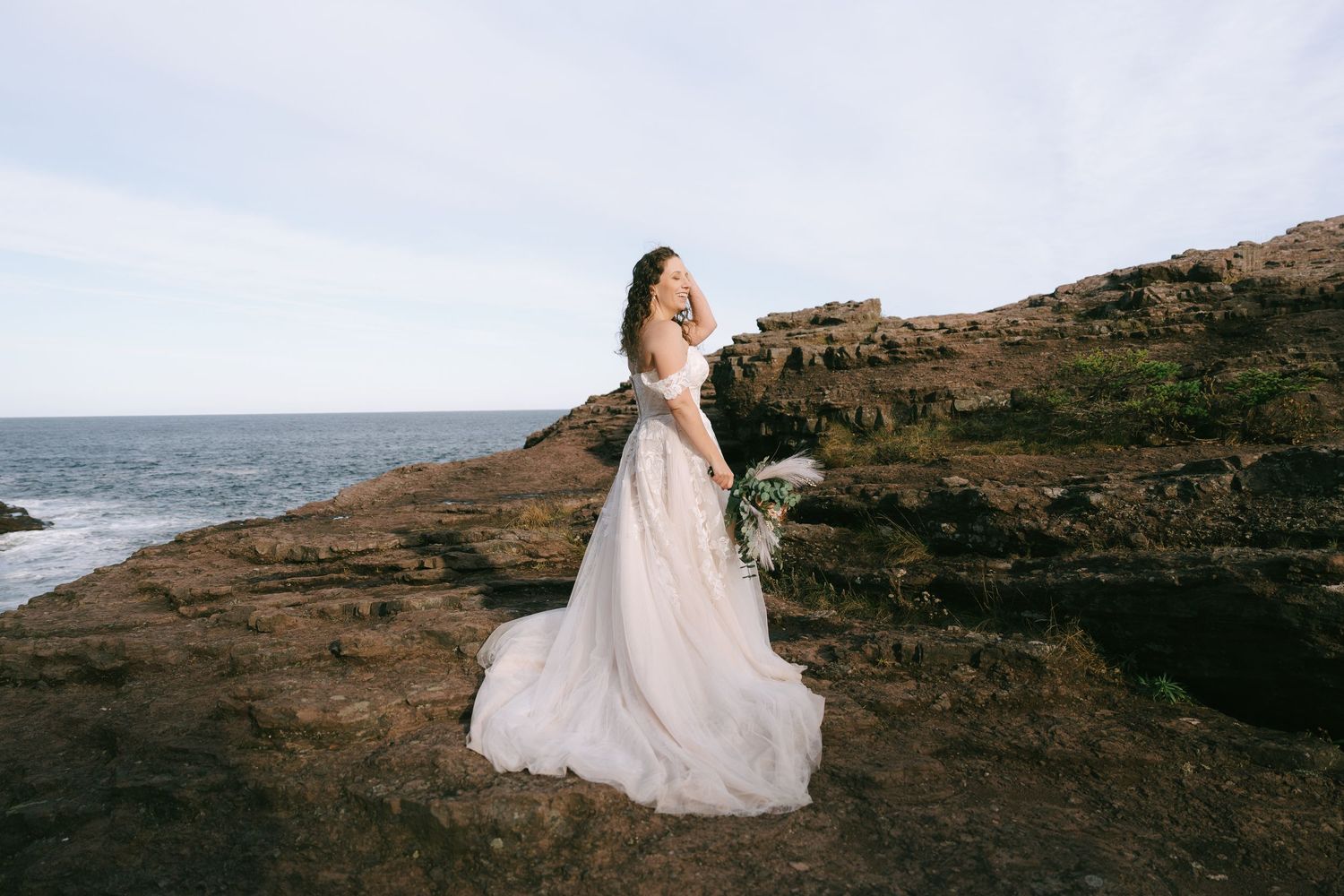 Bride in flowing white gown poses on rocky coastal cliffs at sunset.