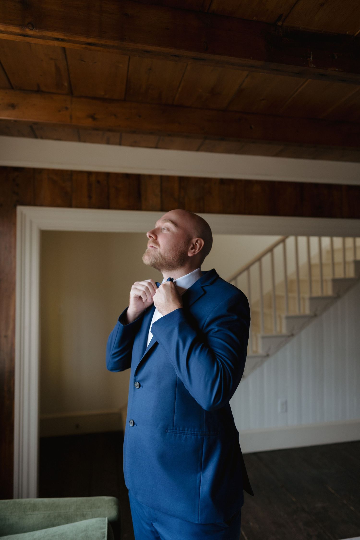 Person in navy blue suit adjusts collar while standing in a room with wooden ceiling beams and natural lighting.
