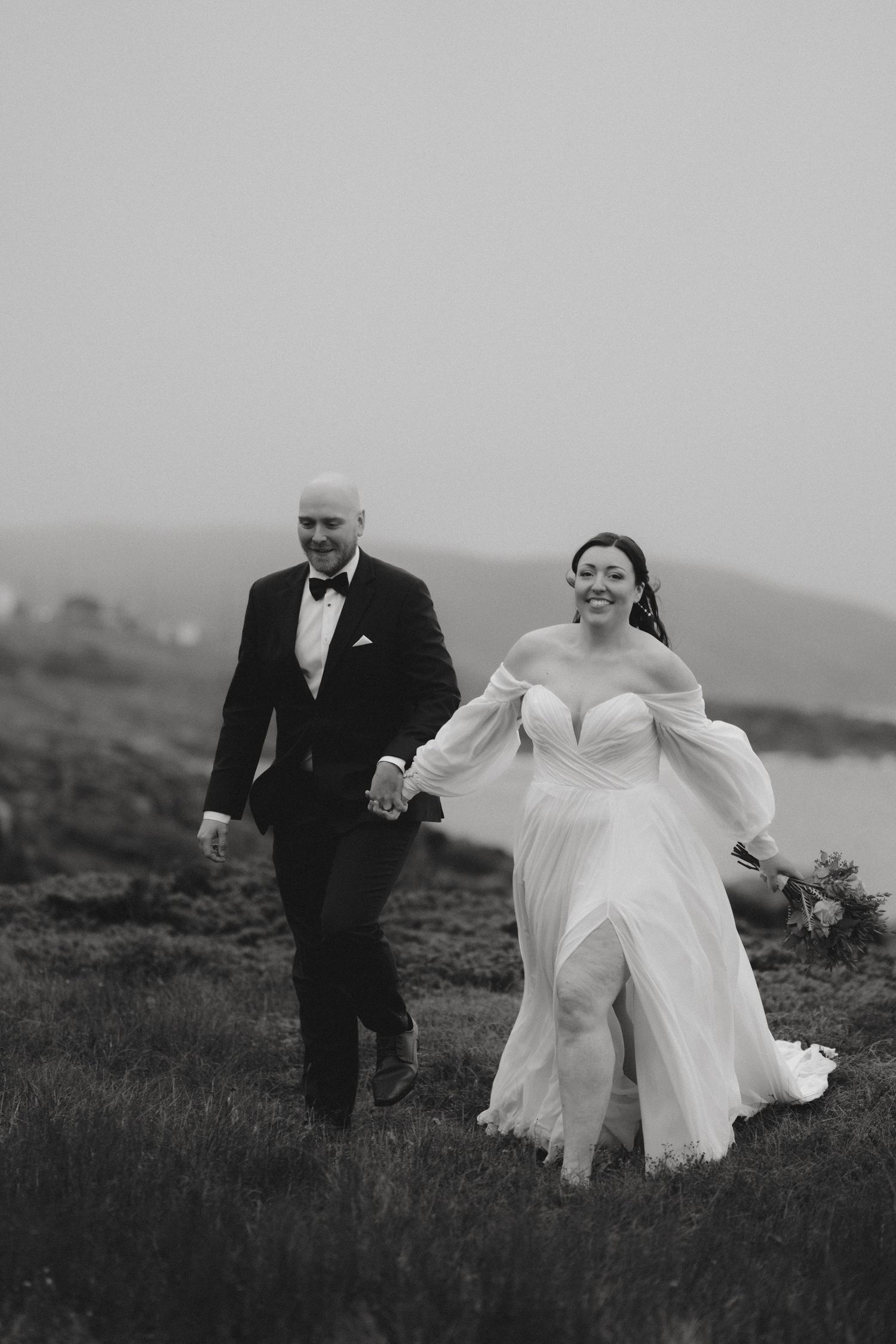 A couple in wedding attire walks hand in hand across a misty hillside in black and white photography.