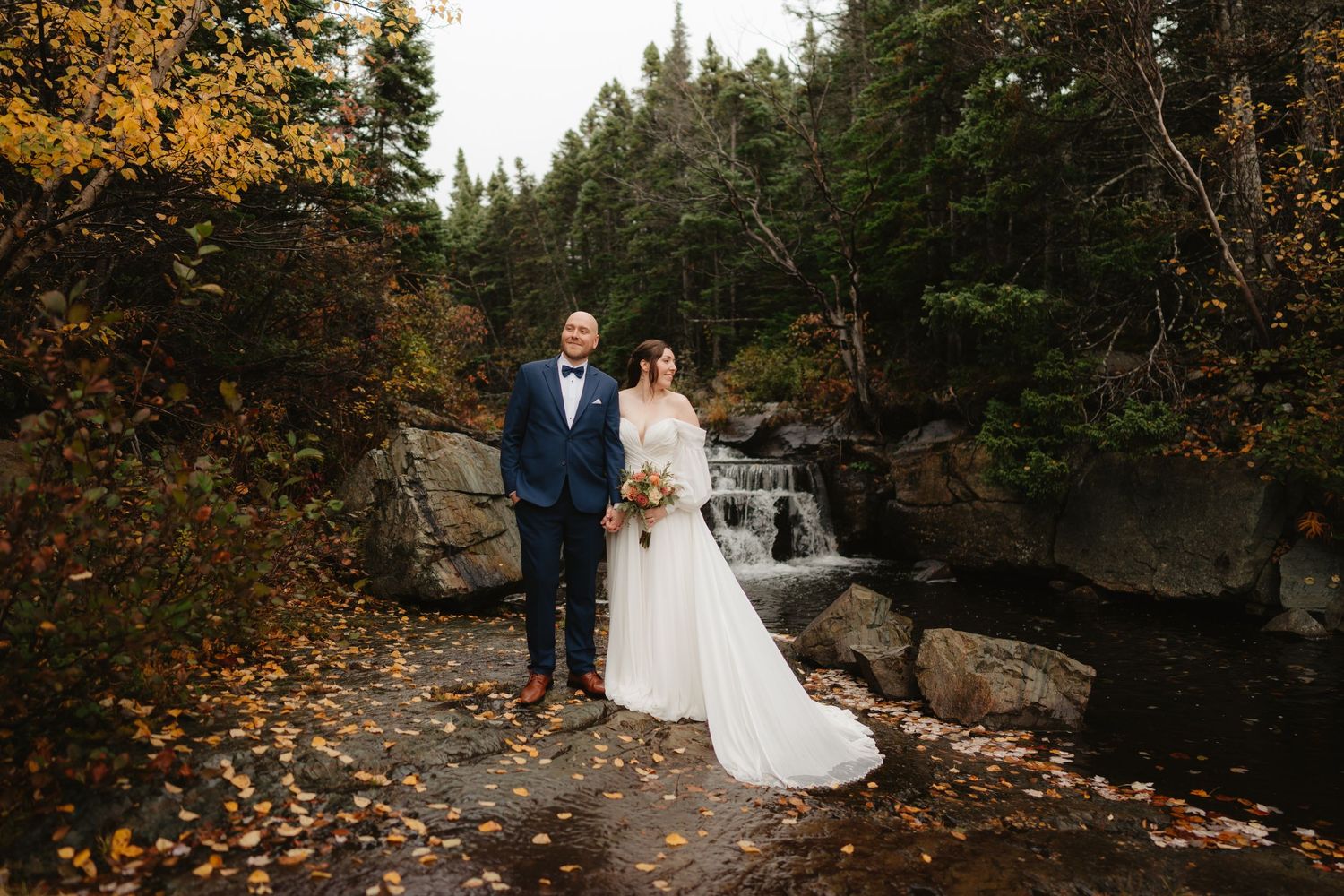 Wedding couple poses near a rushing waterfall in a forest setting with autumn colors and rocky terrain.